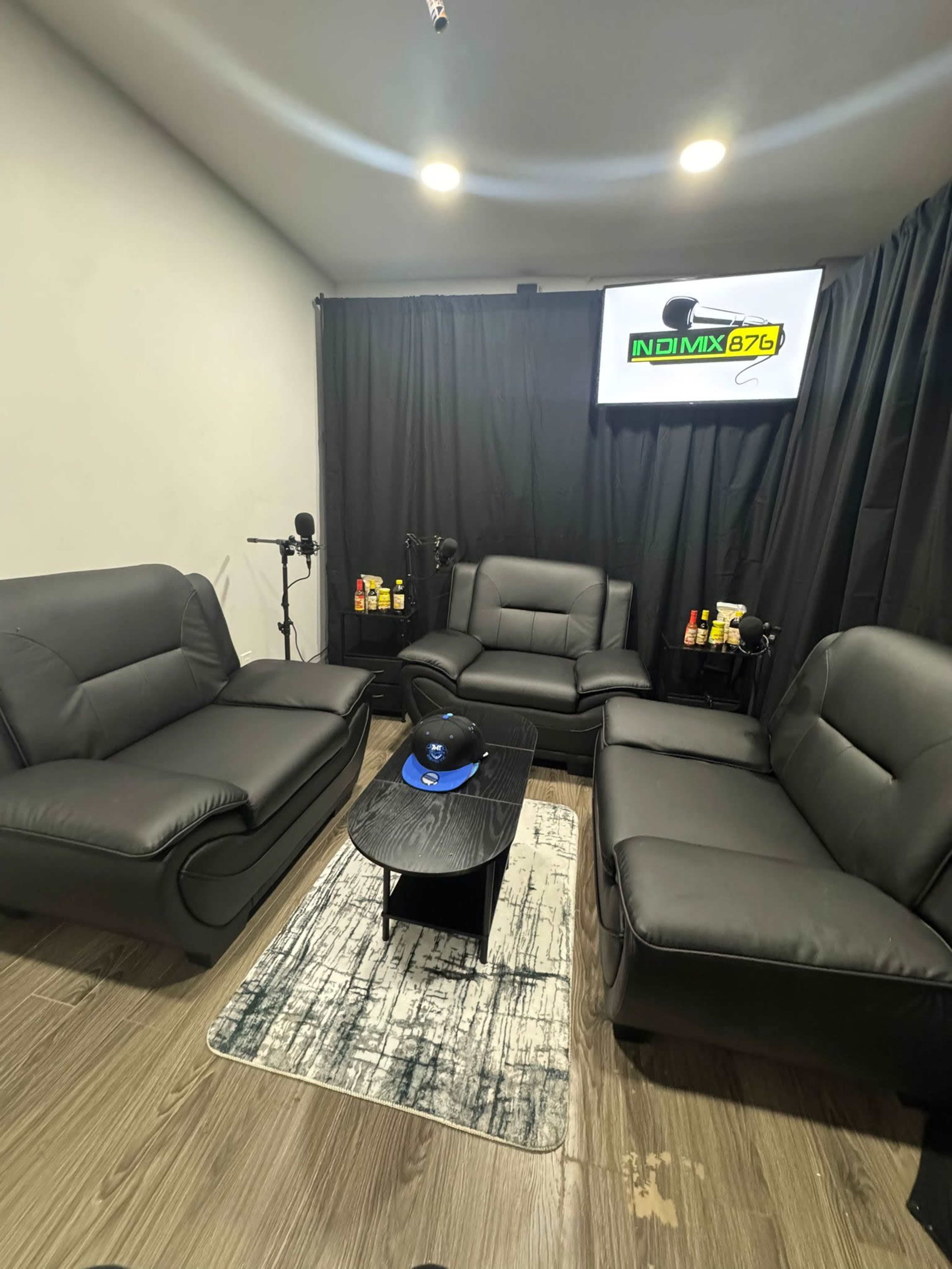 The image shows a cozy lounge area with black leather sofas arranged around a low wooden table, featuring a large screen mounted on the wall and a curtain in the background.