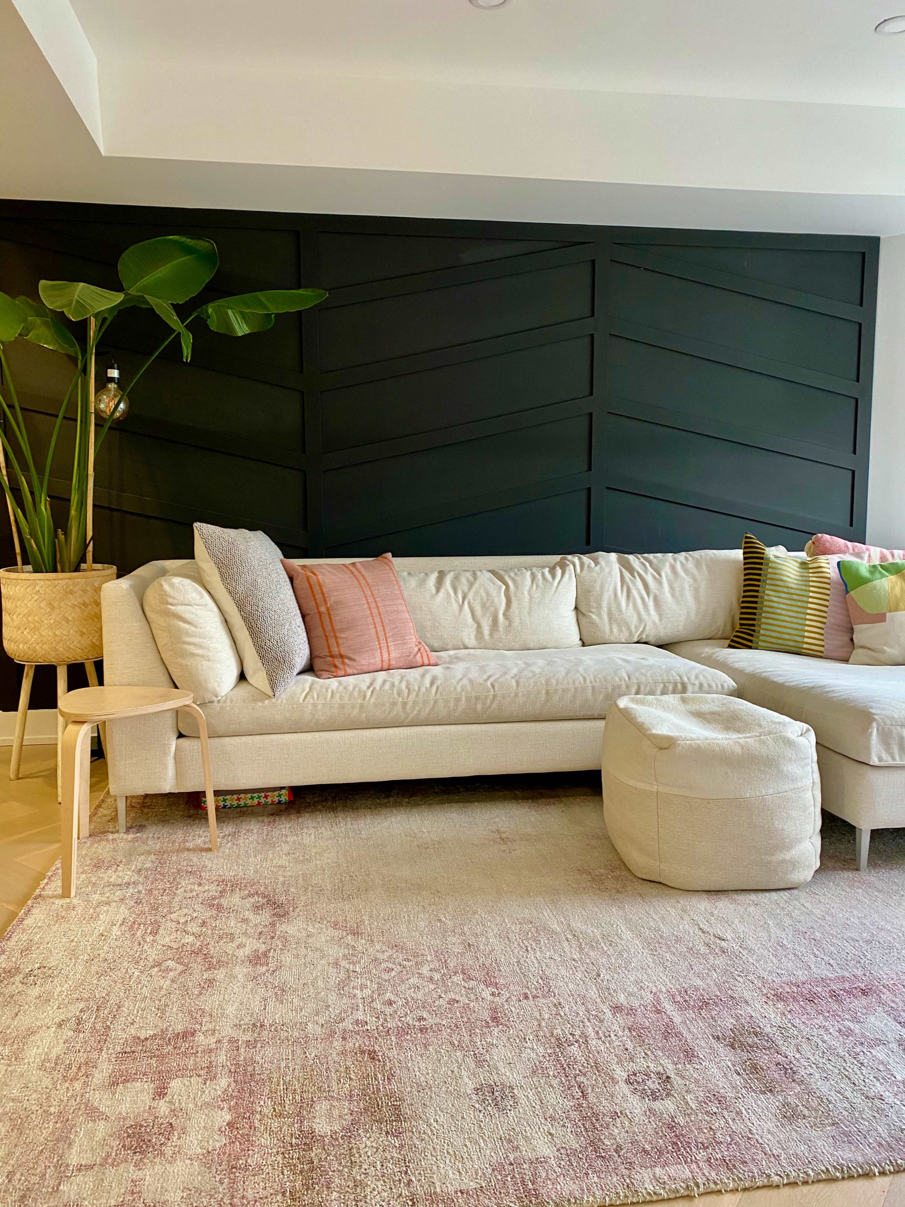 A light-colored sectional sofa adorned with various patterned pillows, placed on a textured rug in a contemporary living room with a dark green accent wall and a large potted plant.