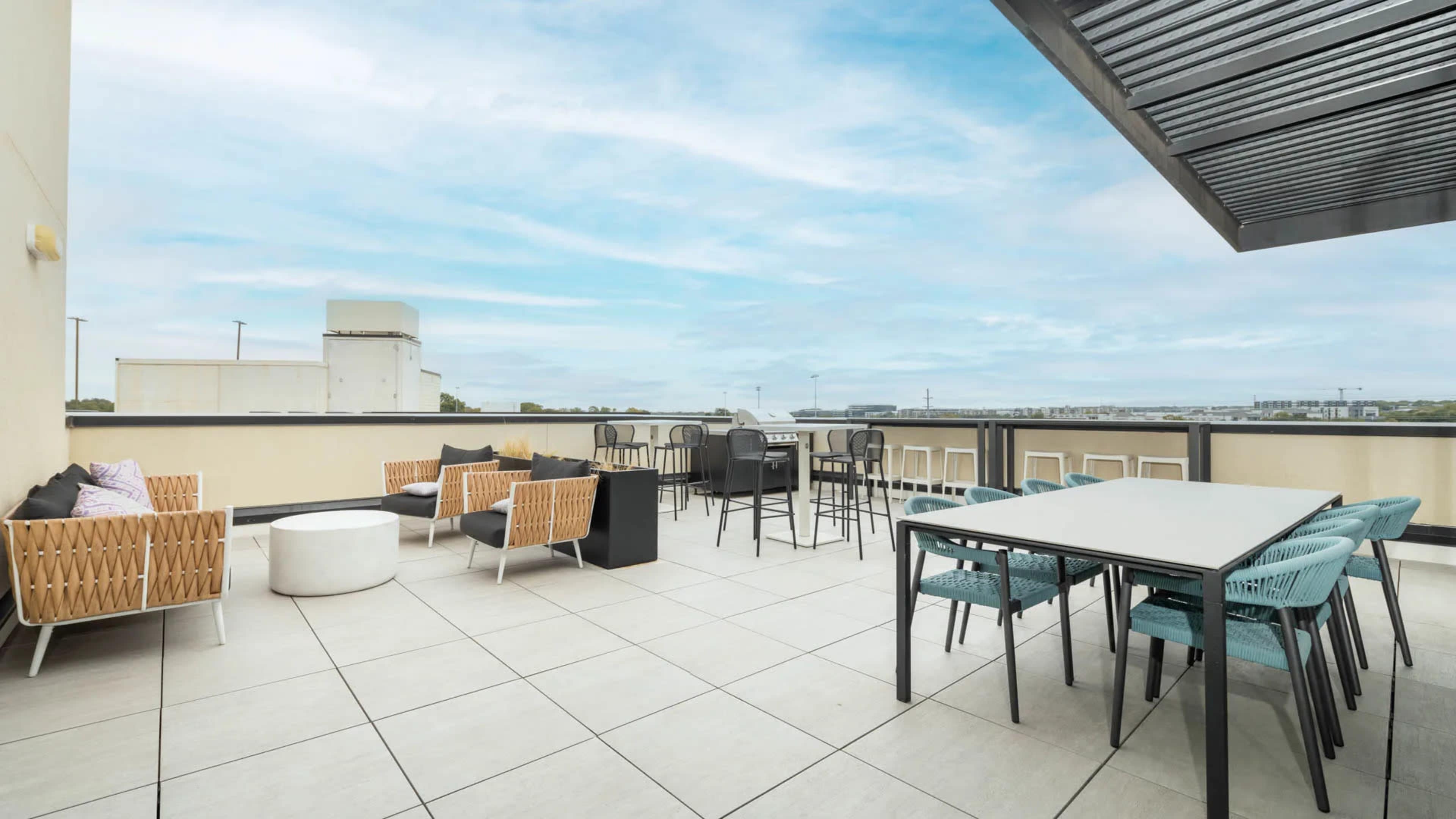 A rooftop terrace featuring seating arrangements with lounge chairs, a large dining table, and a barbecue area under an open sky.