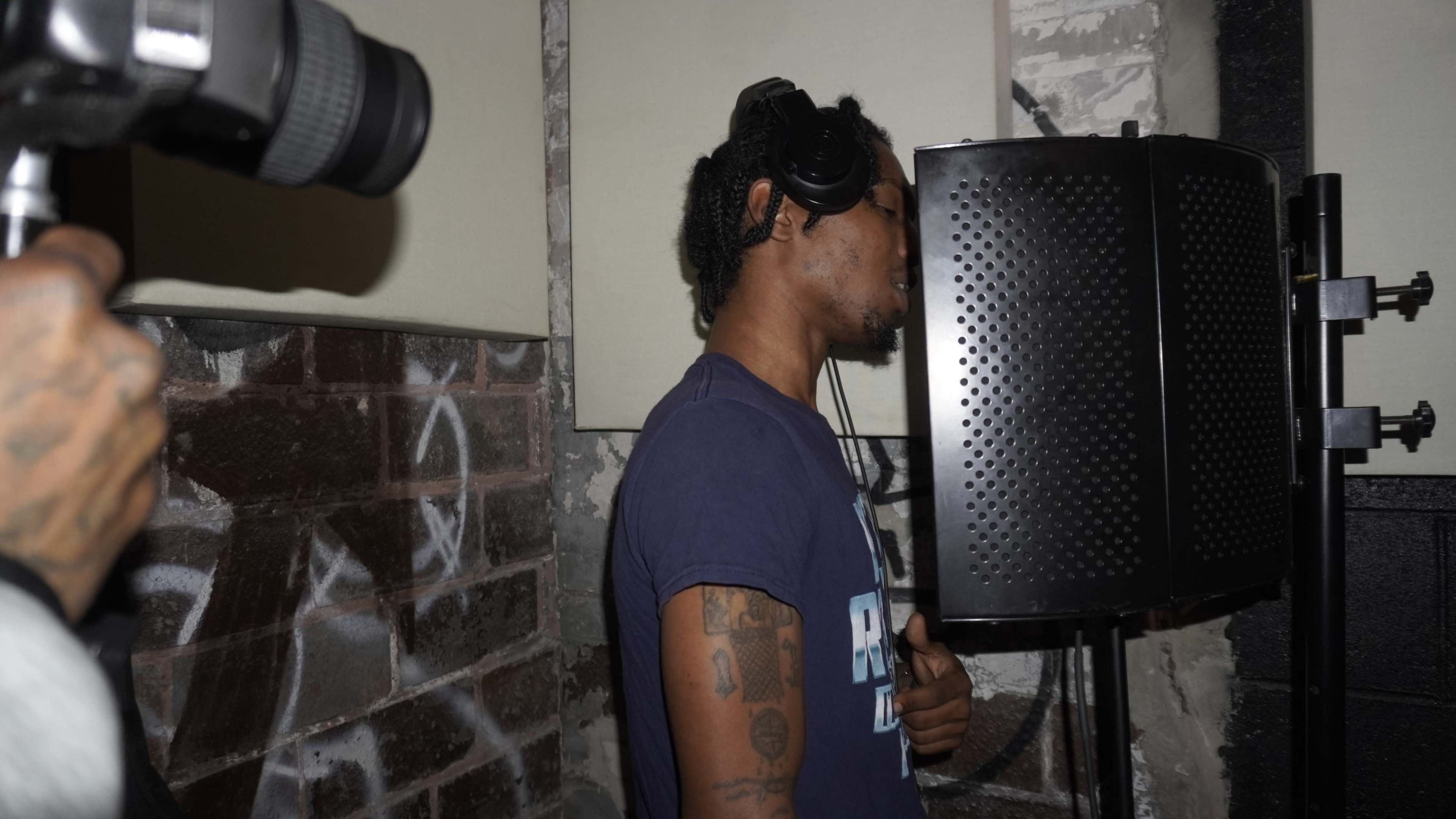 A man wearing headphones is standing in a recording booth while singing or rapping into a microphone.