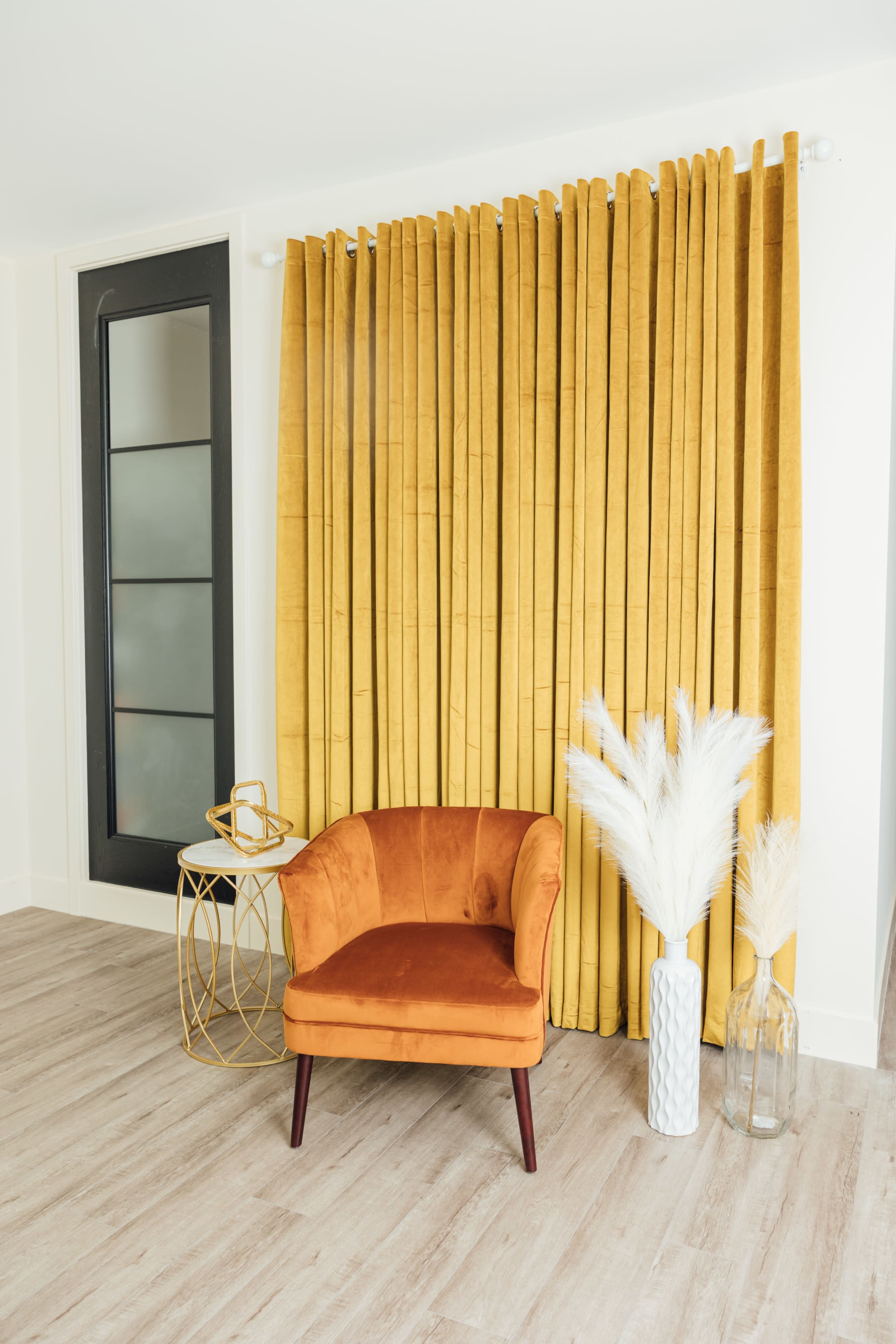 A small orange velvet chair is positioned next to a side table, with long yellow curtains draping behind it and decorative vases nearby.