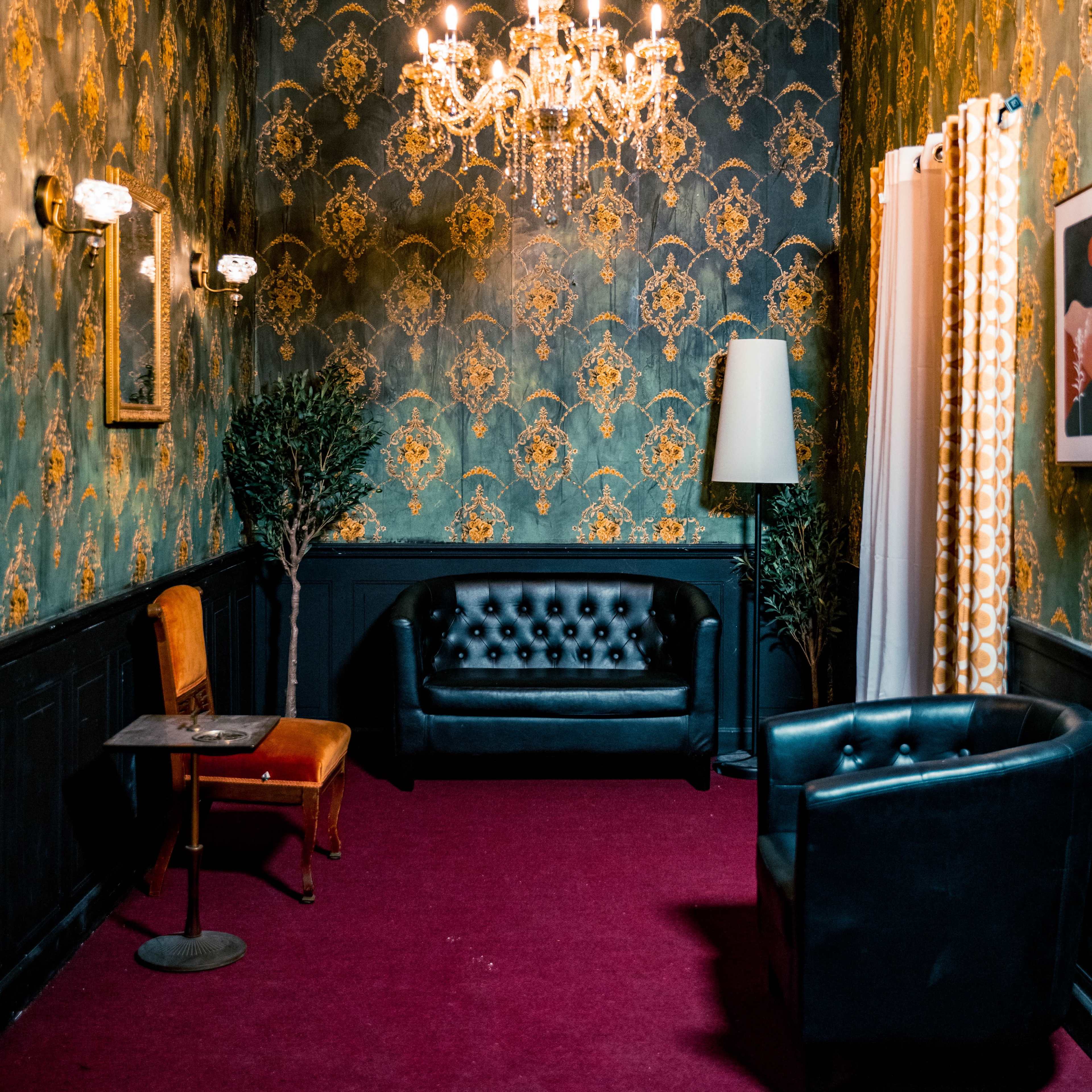 A small, elegantly decorated room featuring a chandelier, vintage wallpaper, and a combination of black leather seating options.