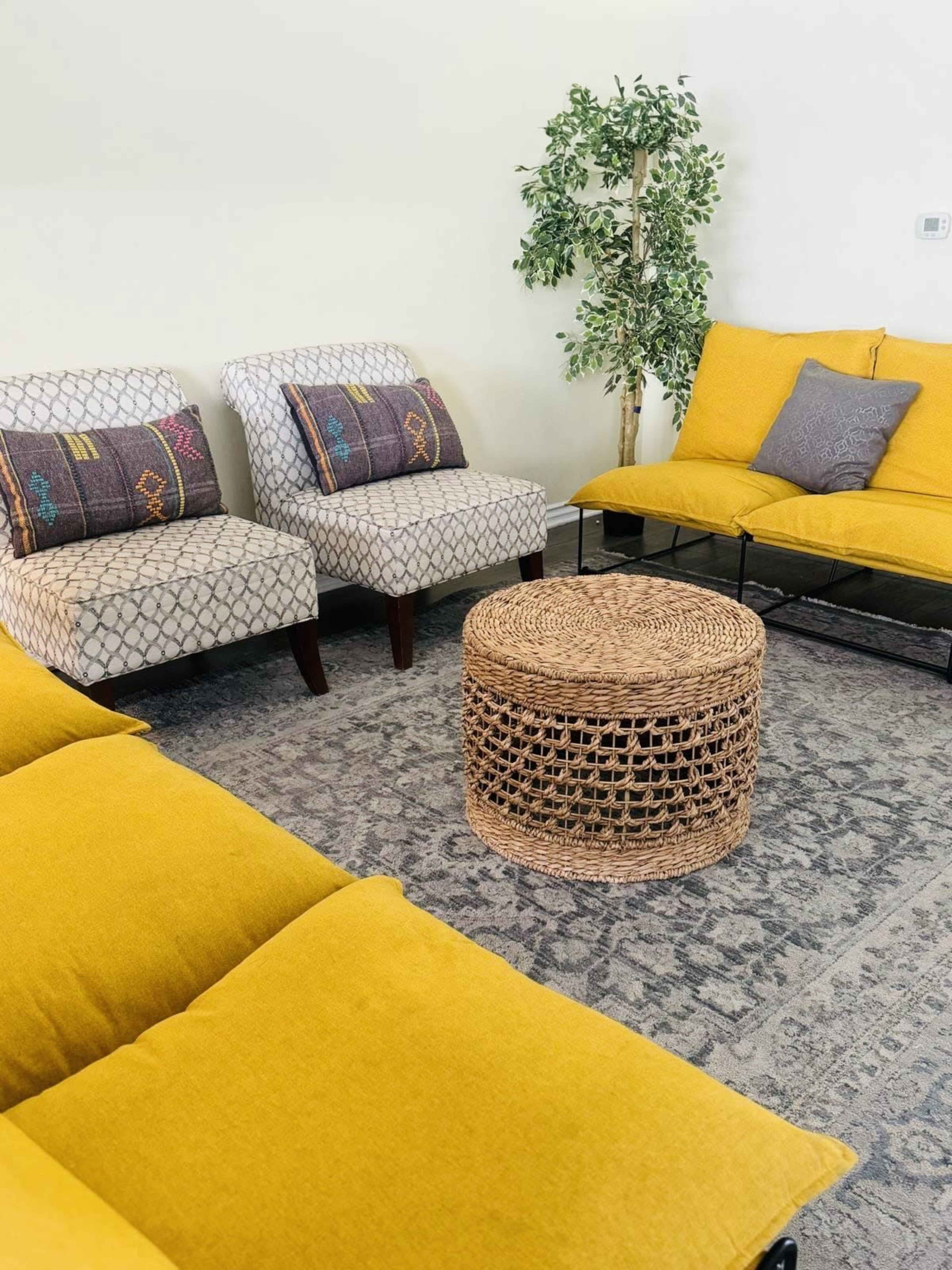 The image shows a living room area featuring a yellow fabric sofa, two patterned armchairs, a circular woven coffee table, and a small decorative plant in the corner.