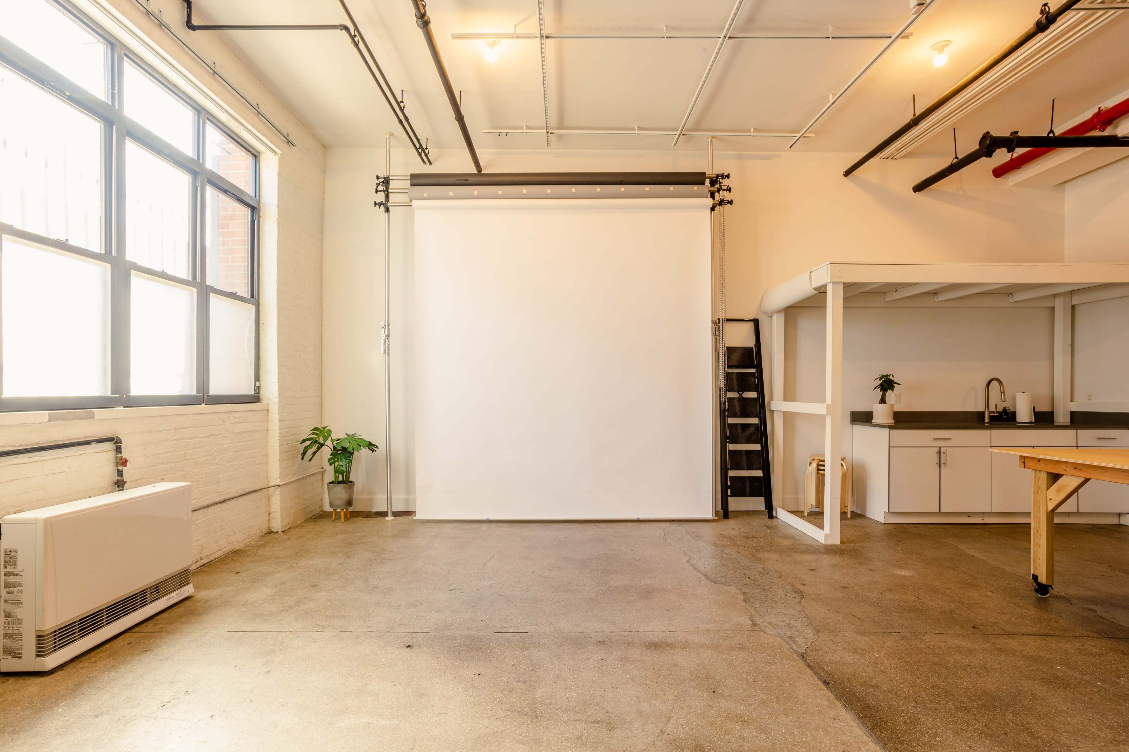 A bright, open studio space featuring a blank white backdrop, a kitchen area, and a ladder against a wall.