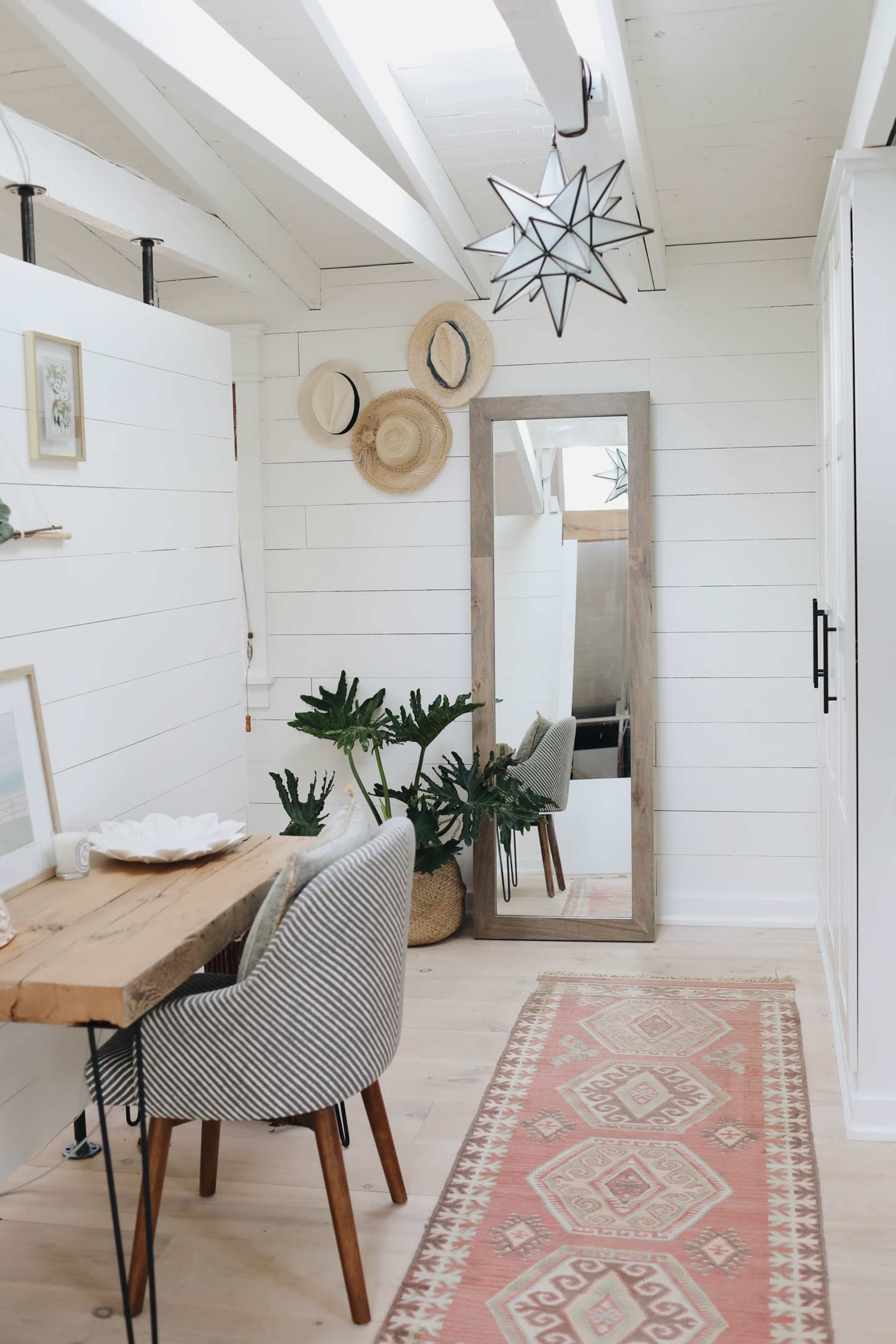 The scene features a bright, modern interior with a wooden dining table and chairs, a large mirror, plants, and decorative wall hangings.