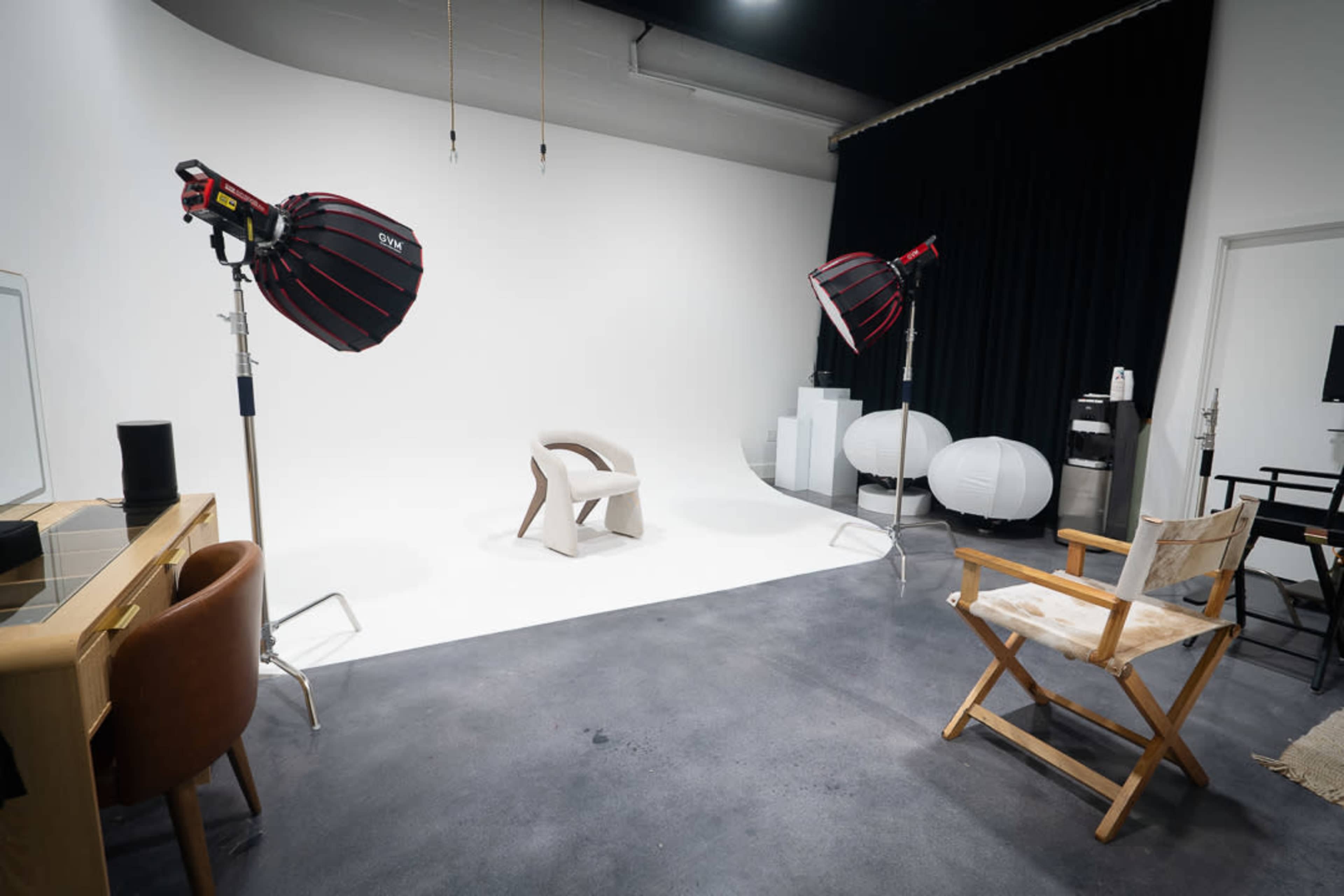The image shows a photography studio featuring two lighting setups, a minimalist chair in the center, and various props including a director's chair and rounded displays.