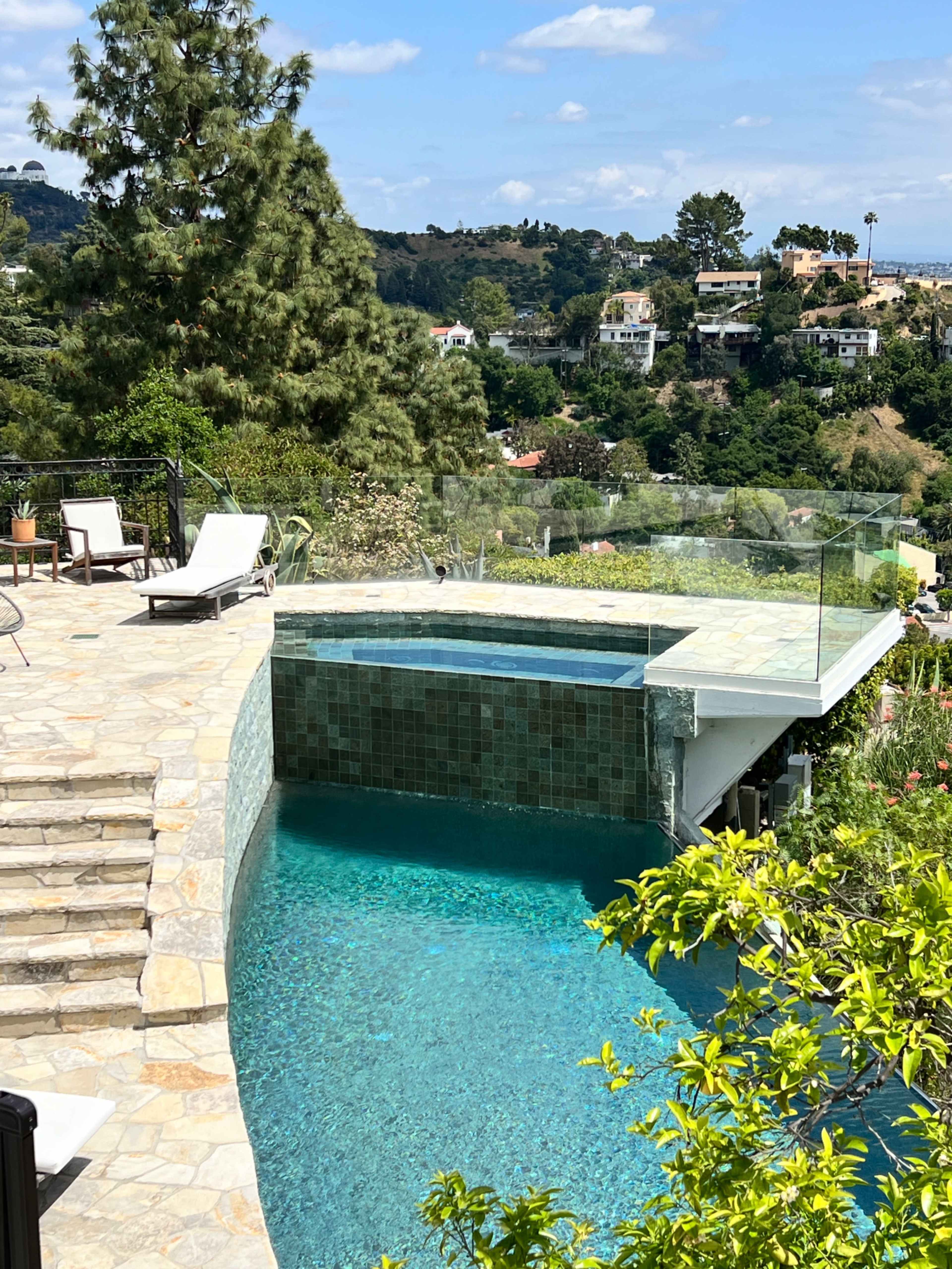The image shows a swimming pool with a glass railing overlooking a hilly landscape and modern houses under a clear blue sky.