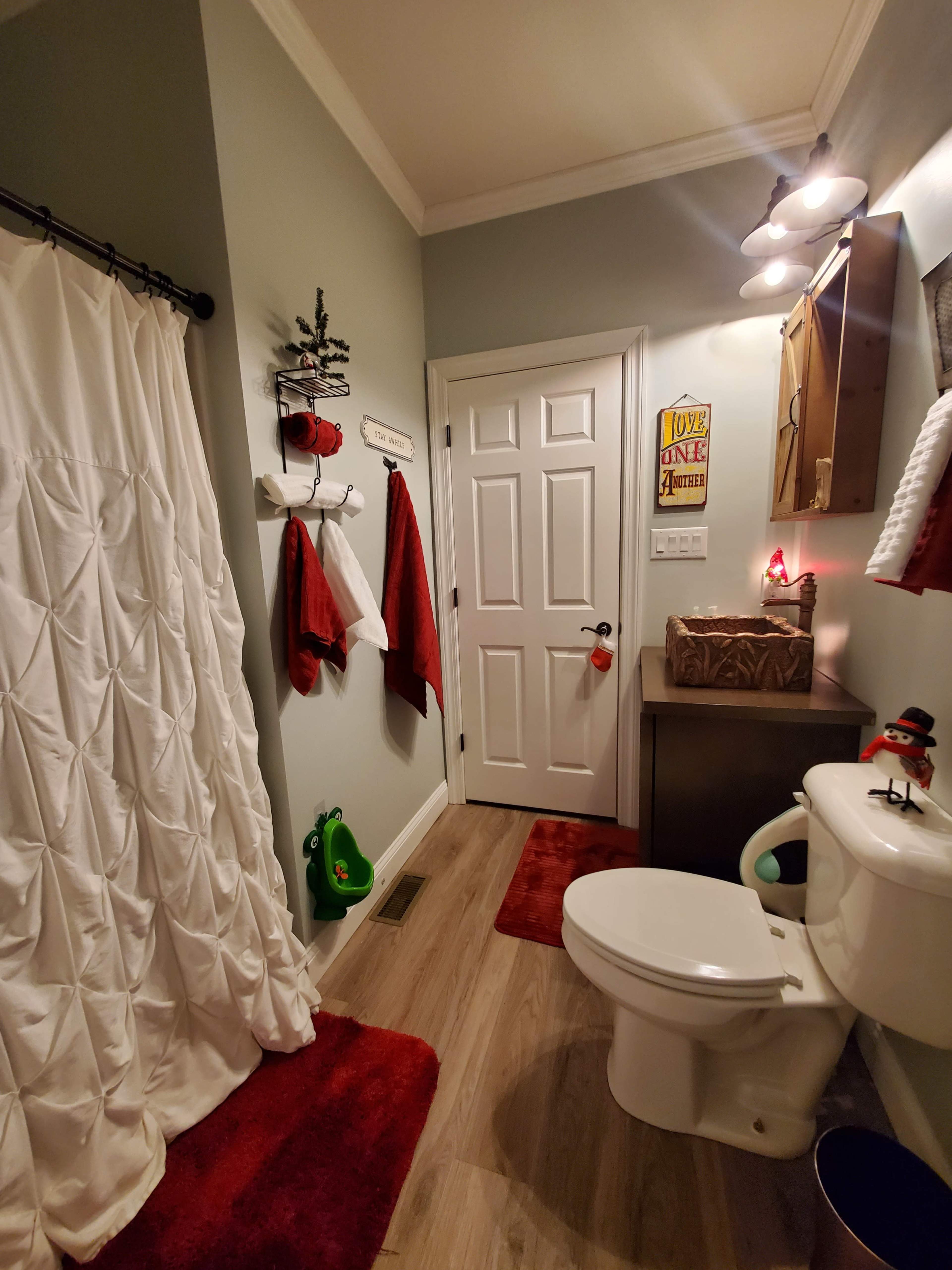 A small bathroom featuring a white shower curtain, a green object on the floor, red towels, and a toilet with a festive snowman decoration.