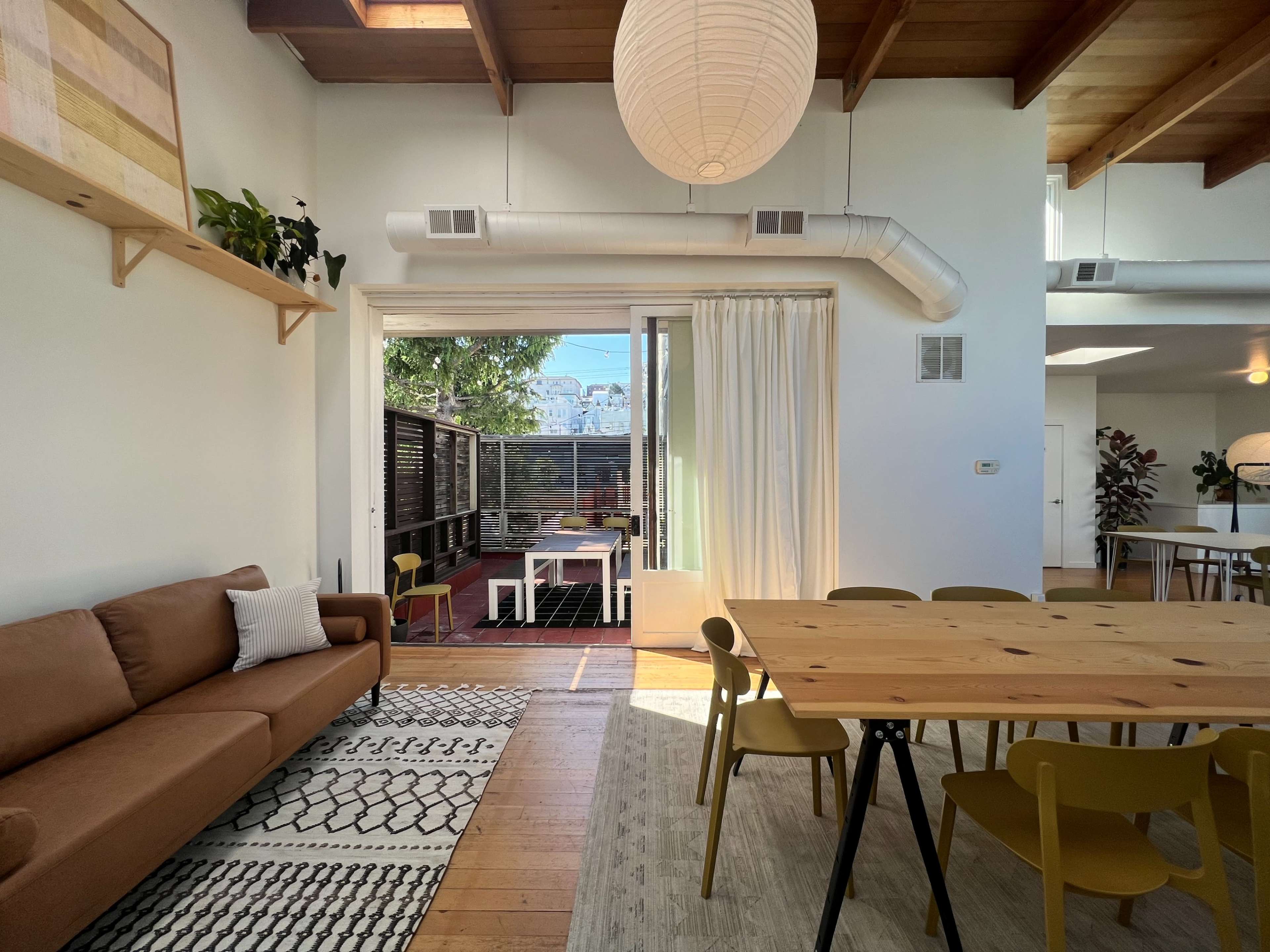 The image shows a bright, open living space featuring a brown sofa, a wooden dining table with yellow chairs, and large windows leading to an outdoor area.