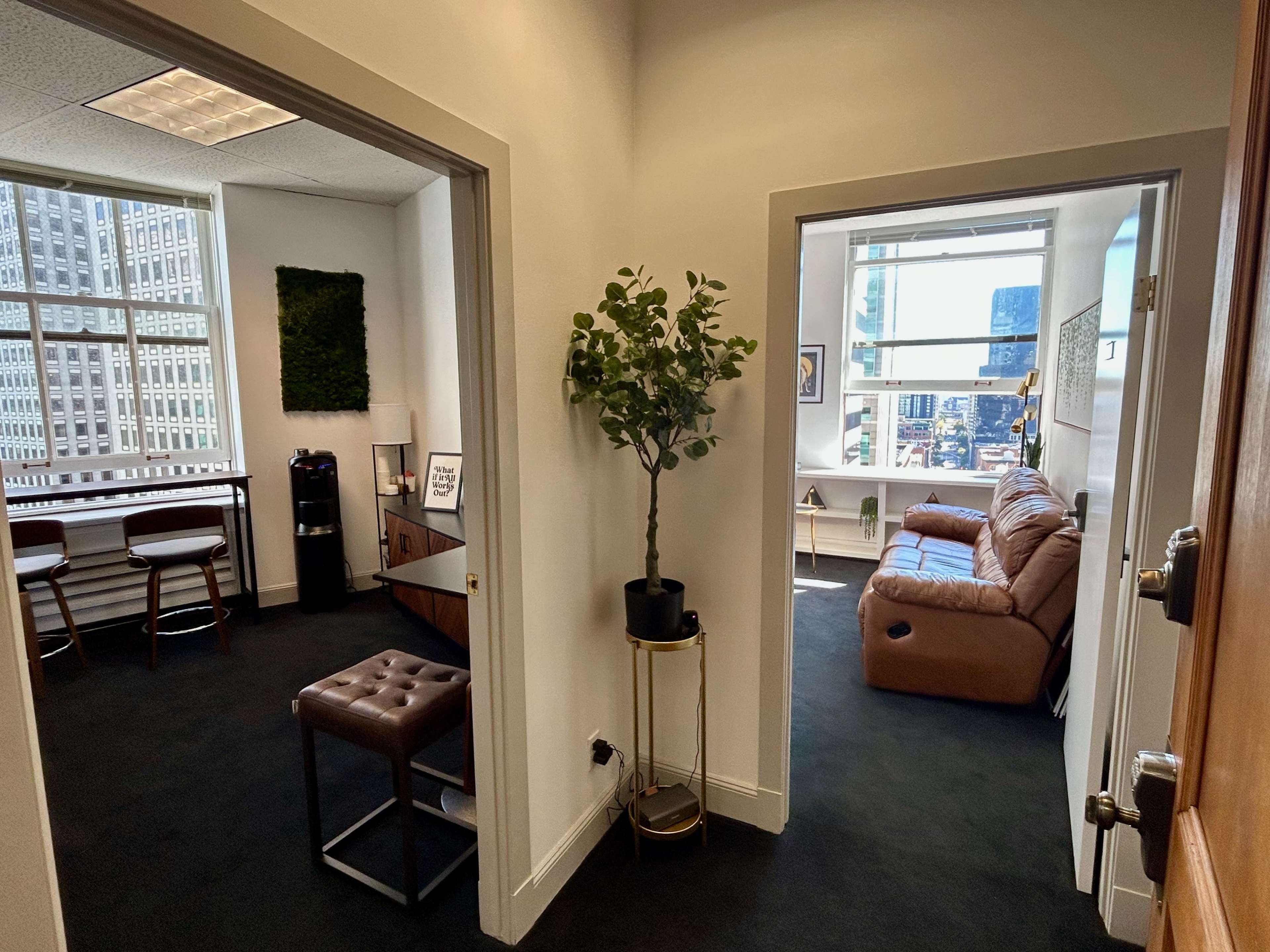A hallway connecting two office spaces, one featuring a small seating area and the other with a bar-style setup and potted plant.