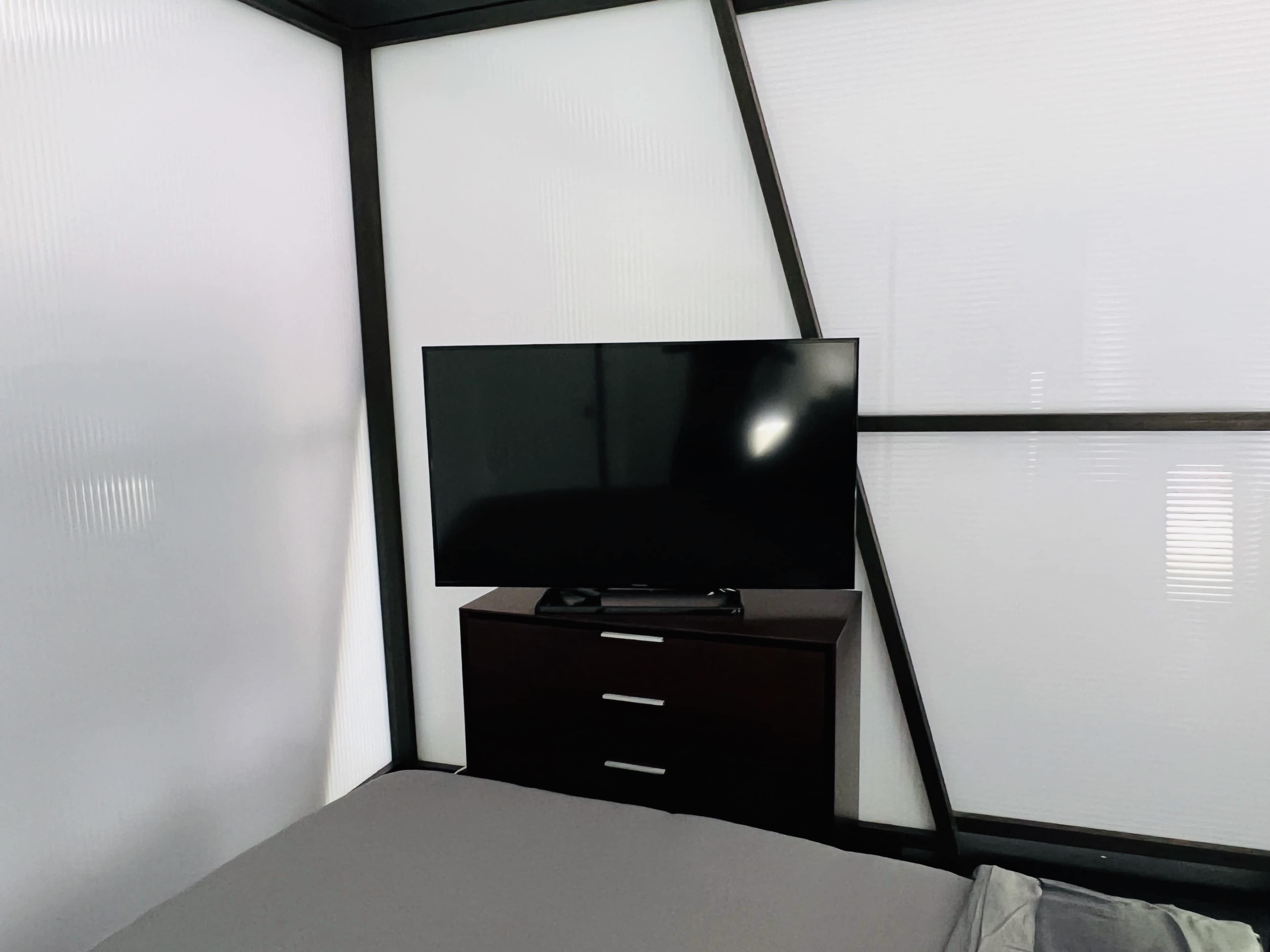 The image shows a minimalist bedroom featuring a bed with a gray cover, a dark wooden dresser, and a flat-screen TV mounted on the wall.