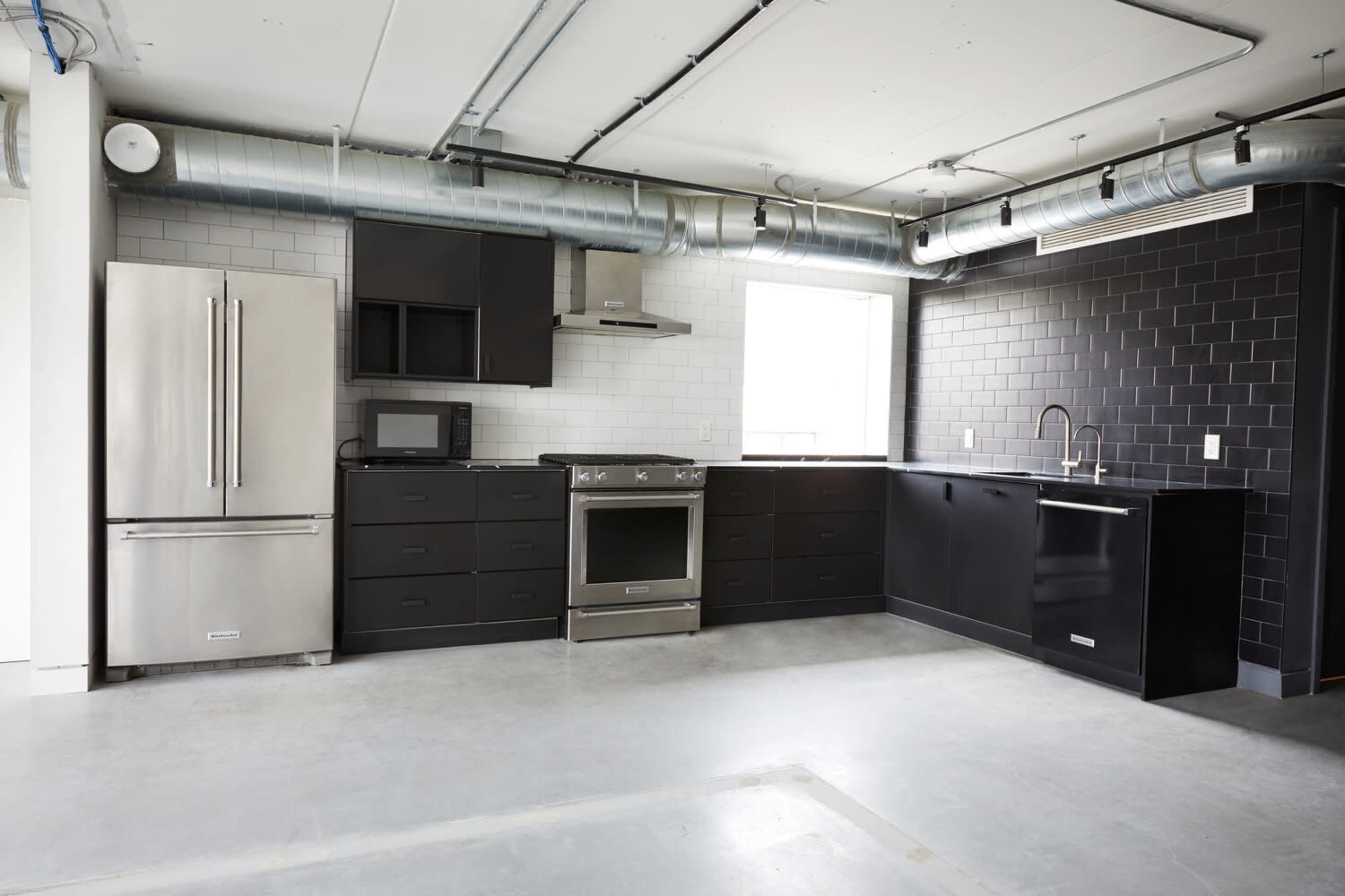 The image shows a modern kitchen featuring stainless steel appliances, black cabinetry, and a minimalist design with exposed ductwork.