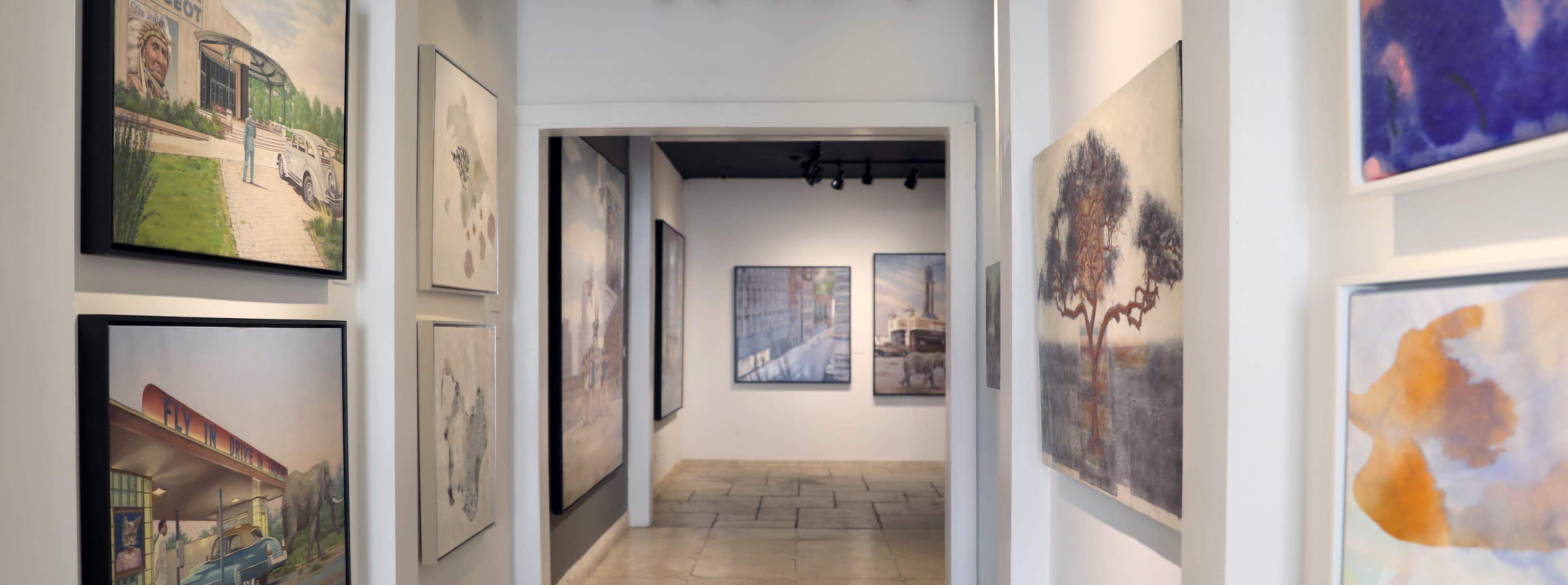 The image shows a gallery hallway lined with various paintings on the walls, leading to a well-lit area in the background.