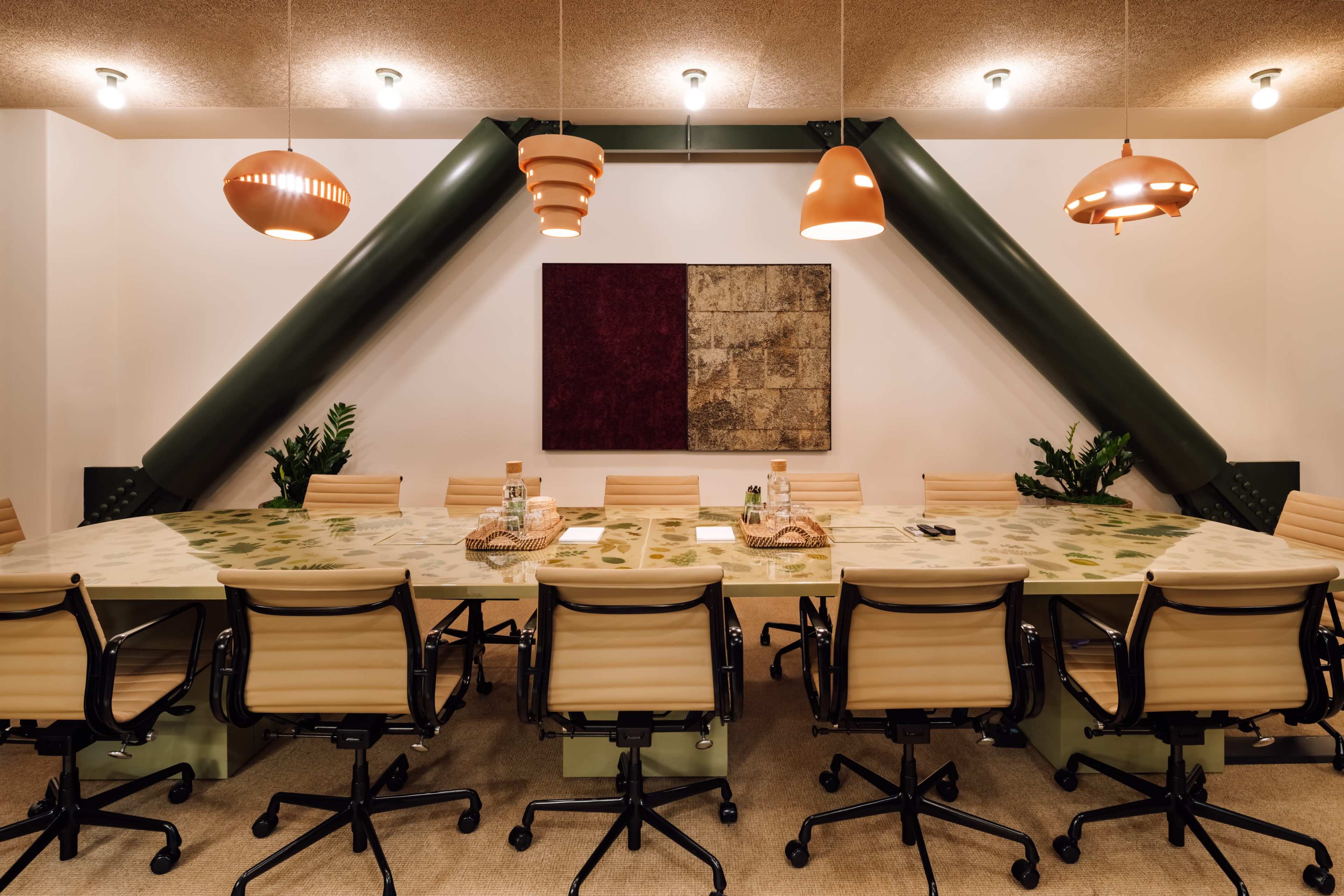 A large conference room features a long table surrounded by beige chairs, with green support beams and pendant lights overhead.