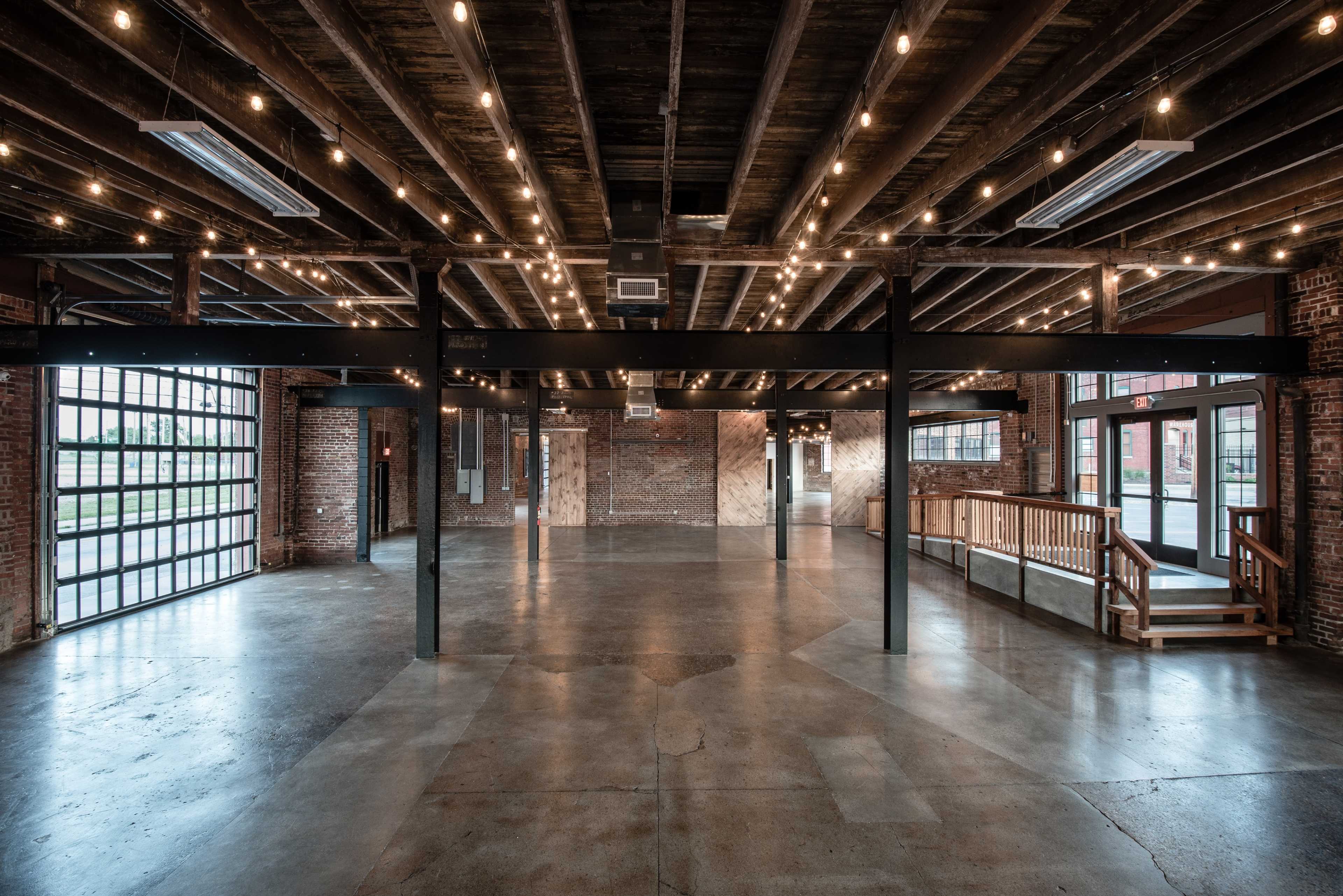 A spacious industrial interior with exposed brick walls, wooden beams, and string lights illuminating the concrete floor.