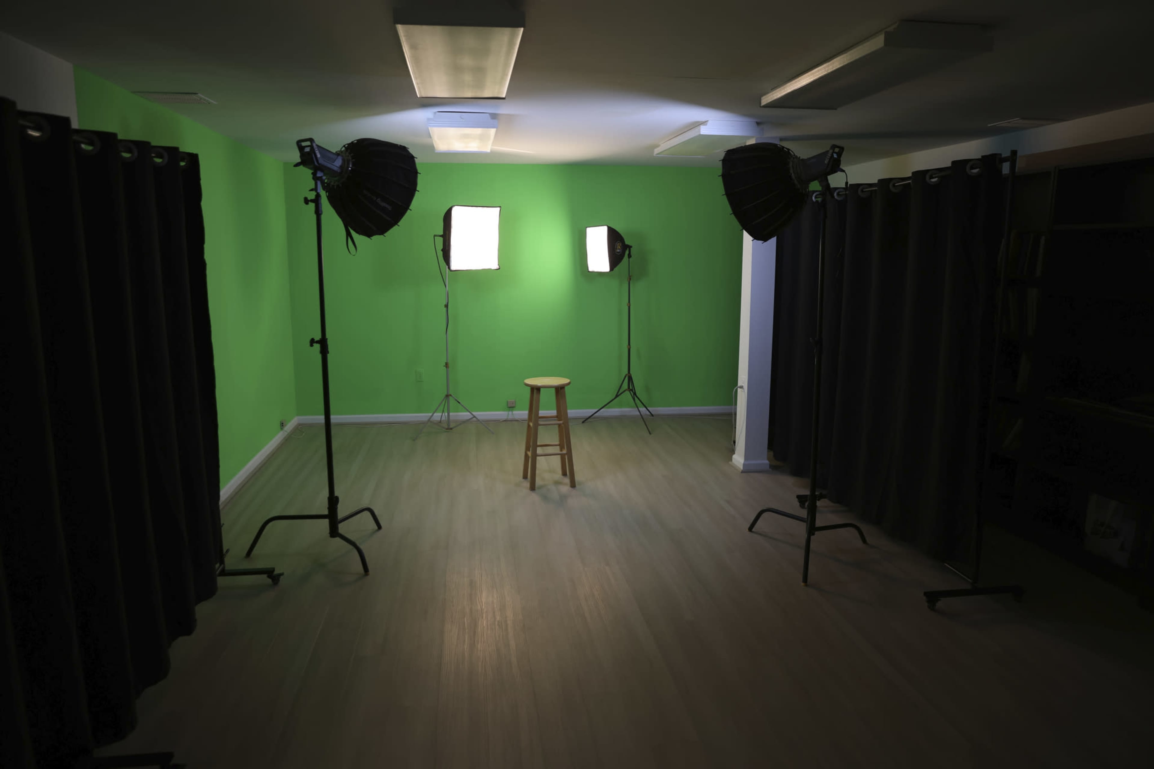 A well-lit studio space features a green backdrop, two softbox lights, a wooden stool in the center, and black curtains at the edges.