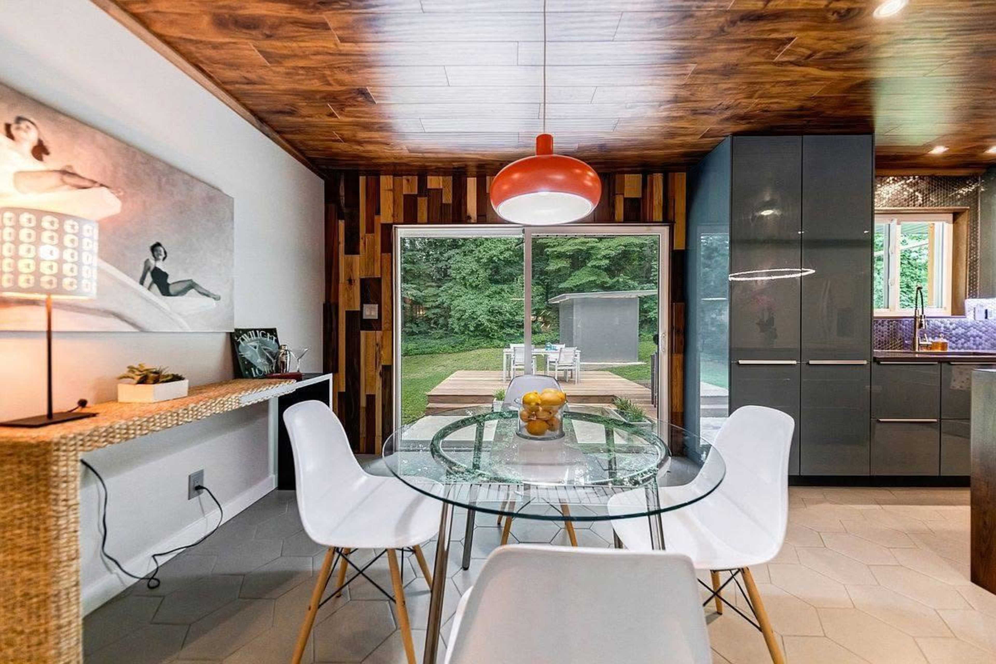 A modern dining area features a round glass table with white chairs, illuminated by a red pendant light, set against a backdrop of wood paneling and a sliding glass door leading to an outdoor deck.