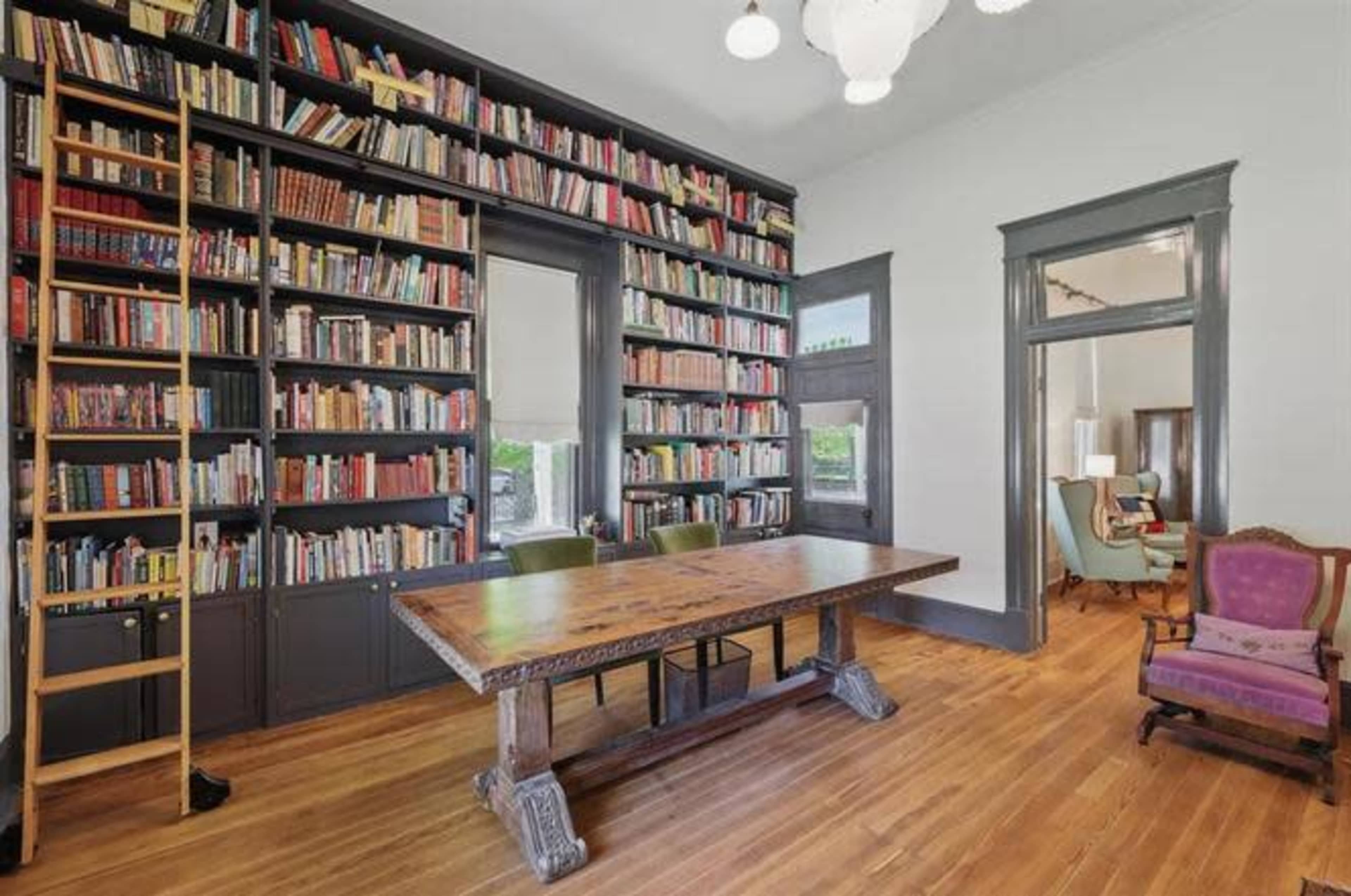 The image shows a library with floor-to-ceiling bookshelves filled with books, a large wooden table in the center, and two green chairs facing the shelves.