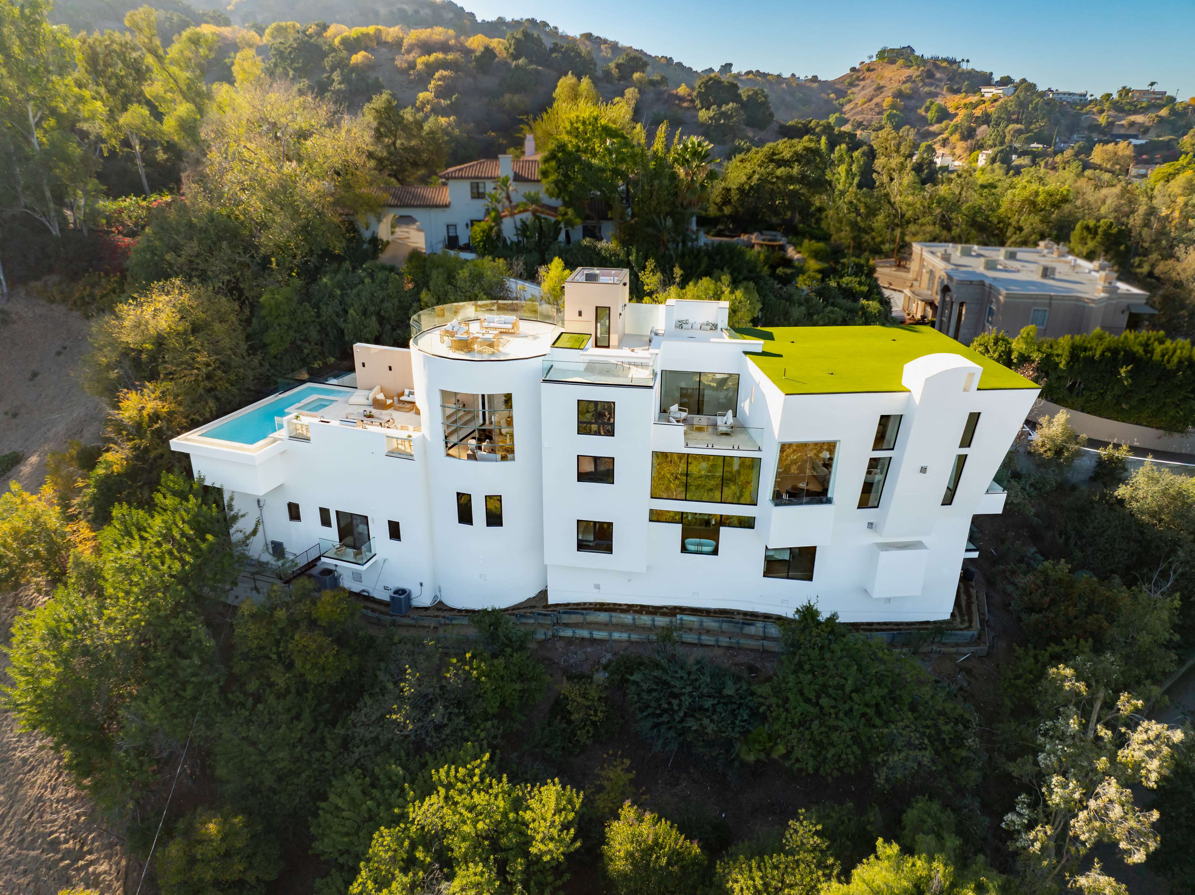 The image shows a modern, multi-level white house with various terraces, a swimming pool, and a green roof, surrounded by trees and a hilly landscape.