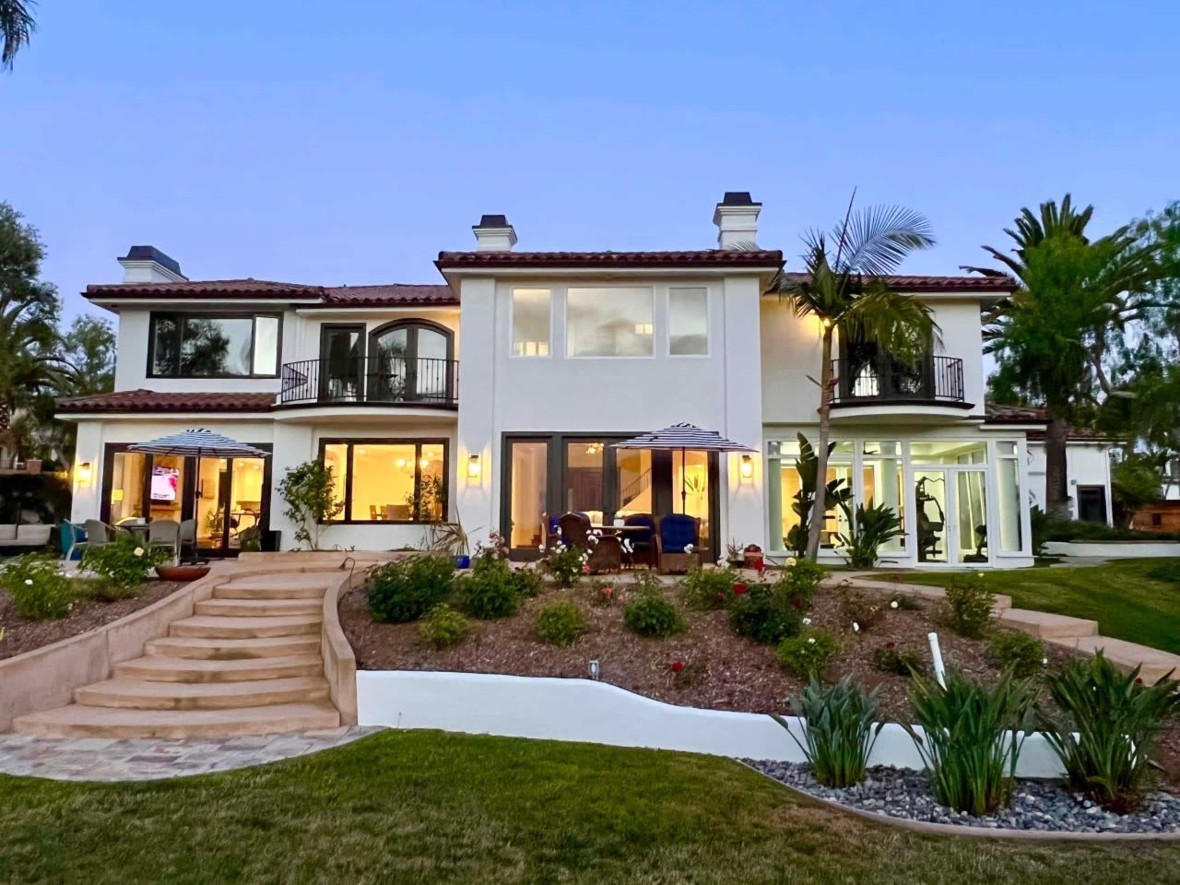 A large, two-story house with a landscaped yard and multiple balconies, illuminated by evening lights.