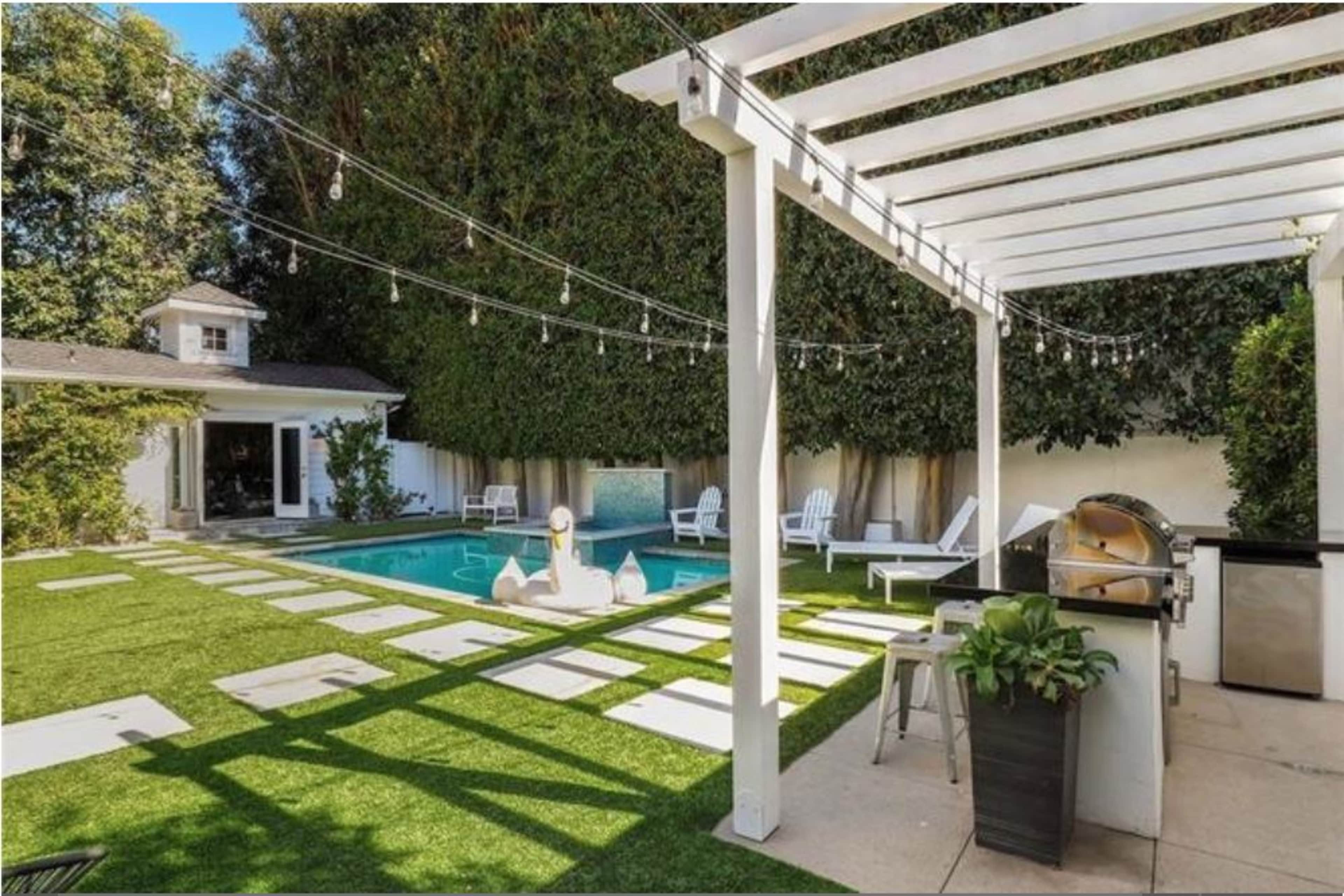 A backyard with a swimming pool, a barbecue area, white lawn chairs, and decorative string lights overhead.