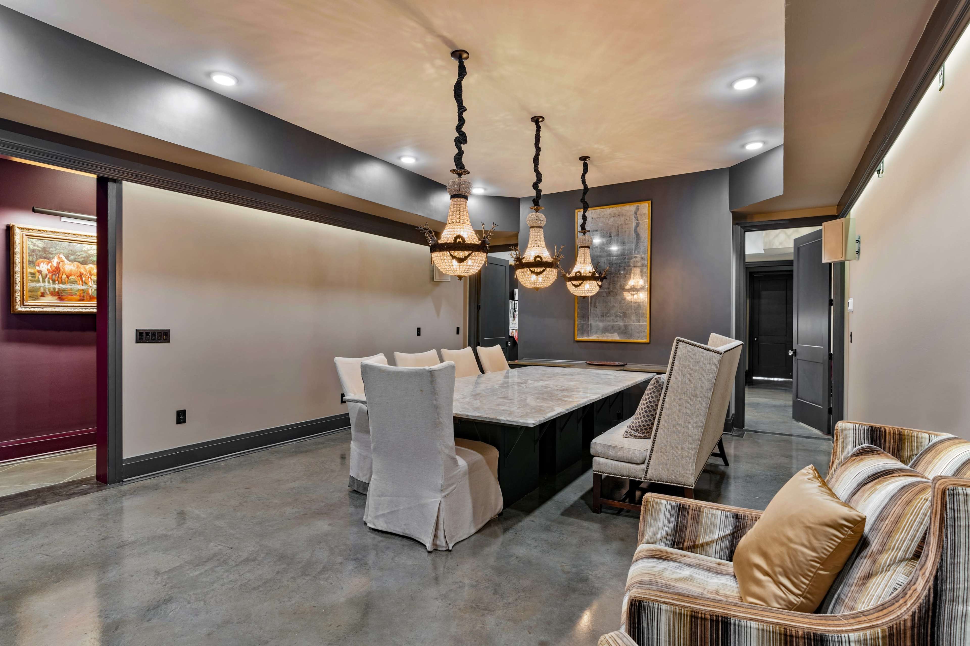 The image shows a spacious dining area featuring a large marble table surrounded by upholstered chairs, illuminated by several hanging light fixtures, with a polished concrete floor and a mix of wall colors.
