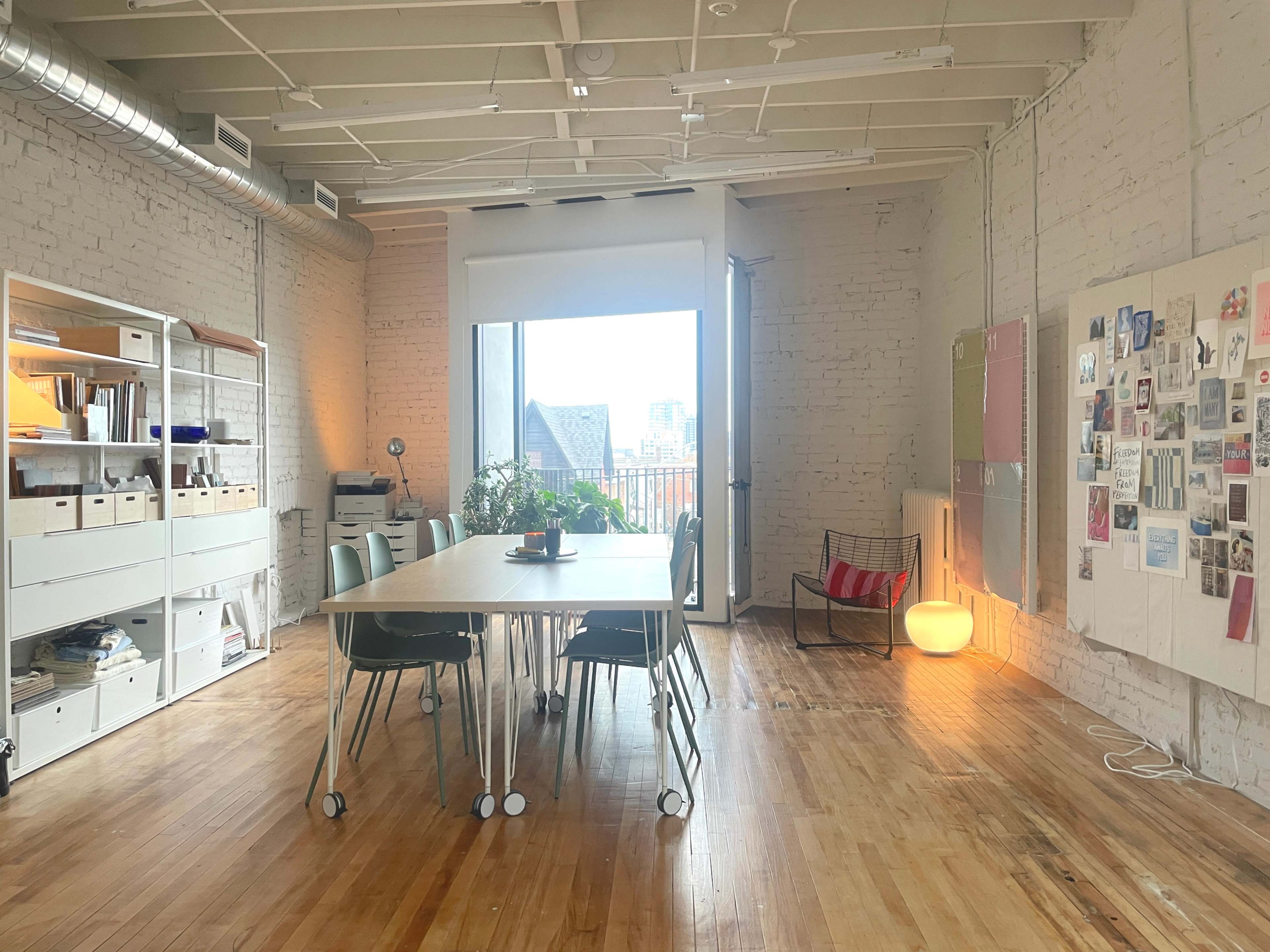 A spacious, well-lit room features a large table with chairs, a storage unit, and a wall covered in various pinned images and documents.