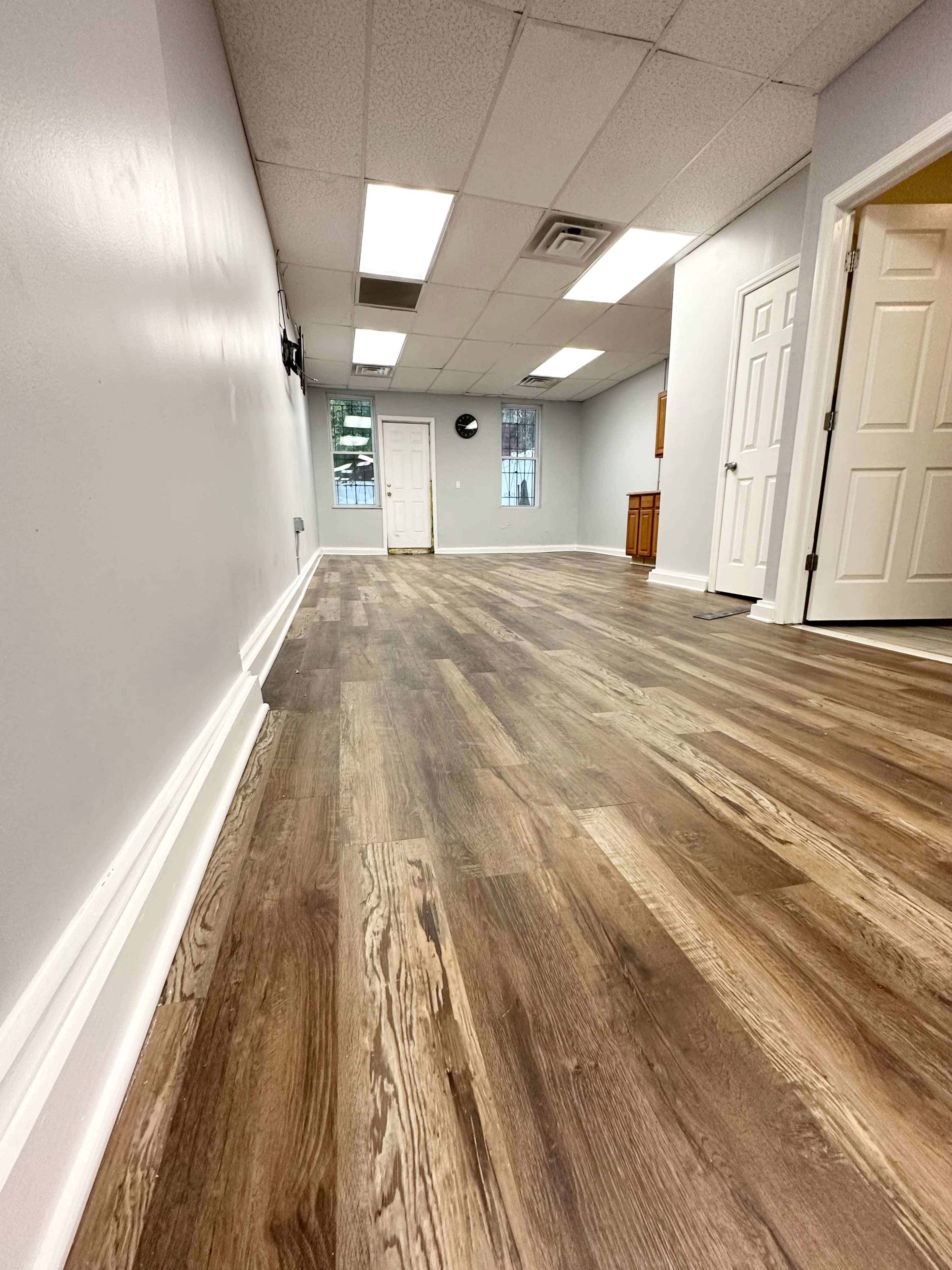 The image shows a spacious, light-colored room with a wooden floor, featuring a closed door and white walls.