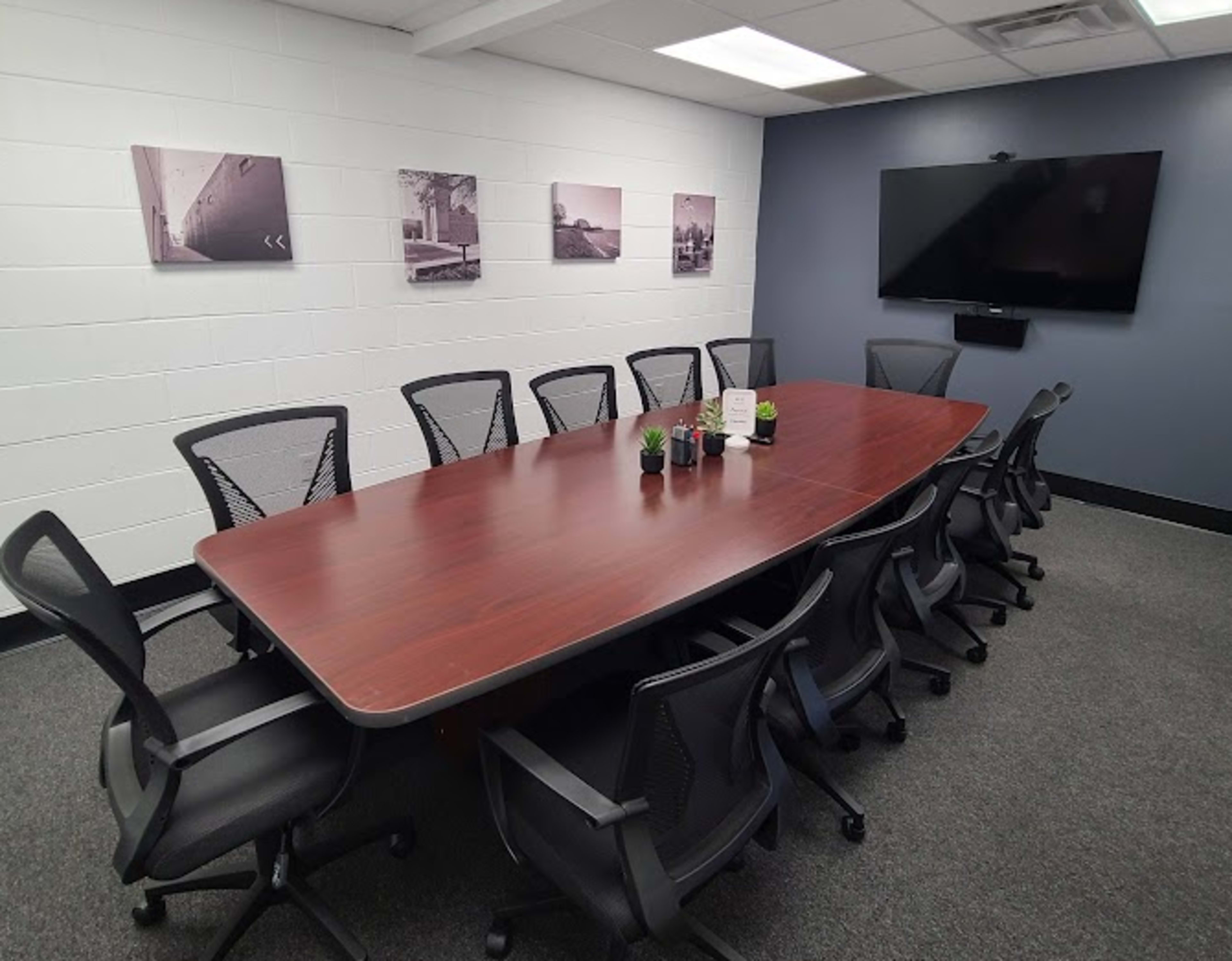 A conference room features a large rectangular wooden table surrounded by black ergonomic chairs, with framed photographs on the walls and a flat-screen TV mounted at one end.