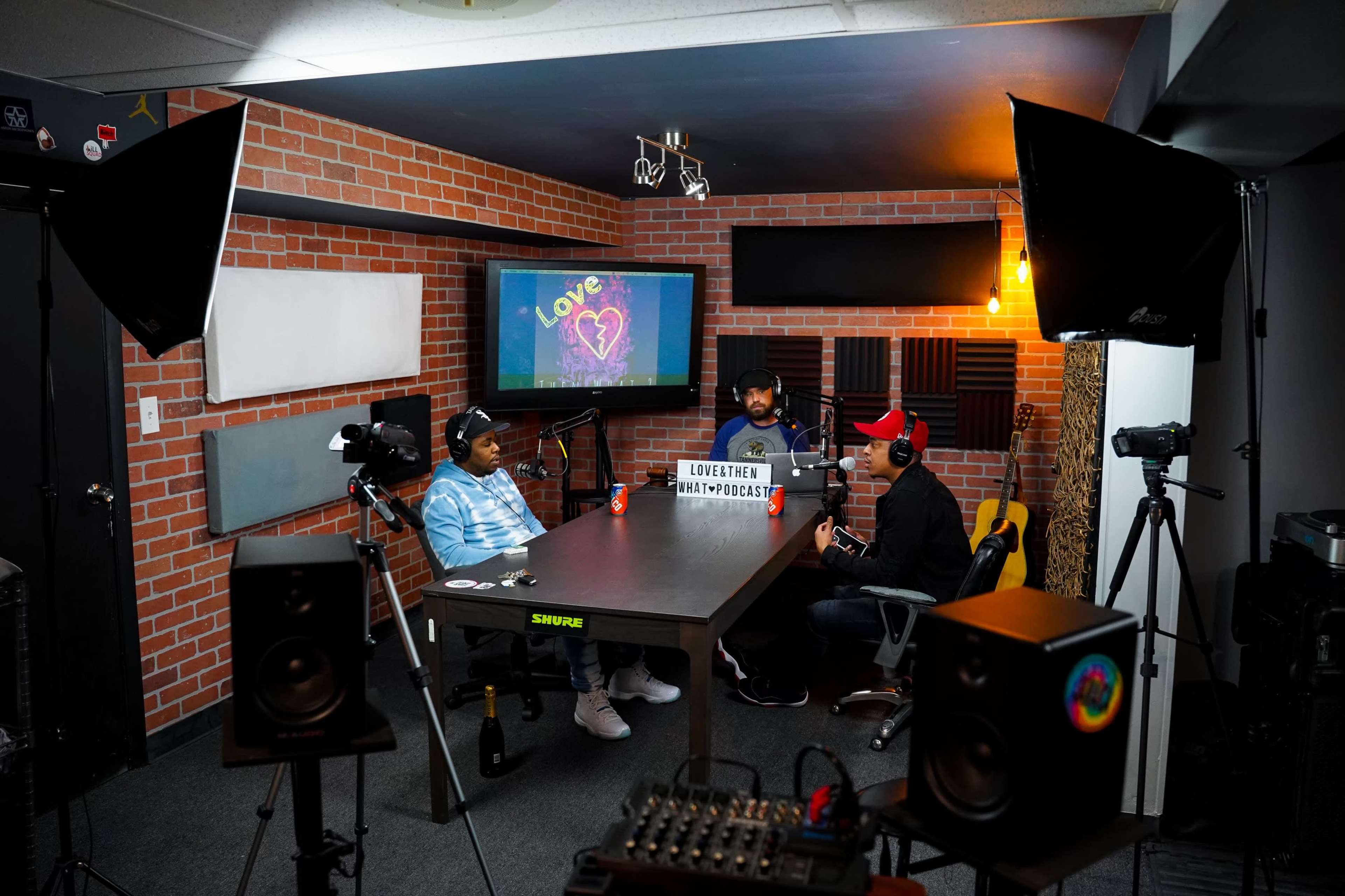 Three individuals are seated at a table in a podcast studio, equipped with microphones and cameras, discussing in front of a monitor displaying a colorful heart graphic.