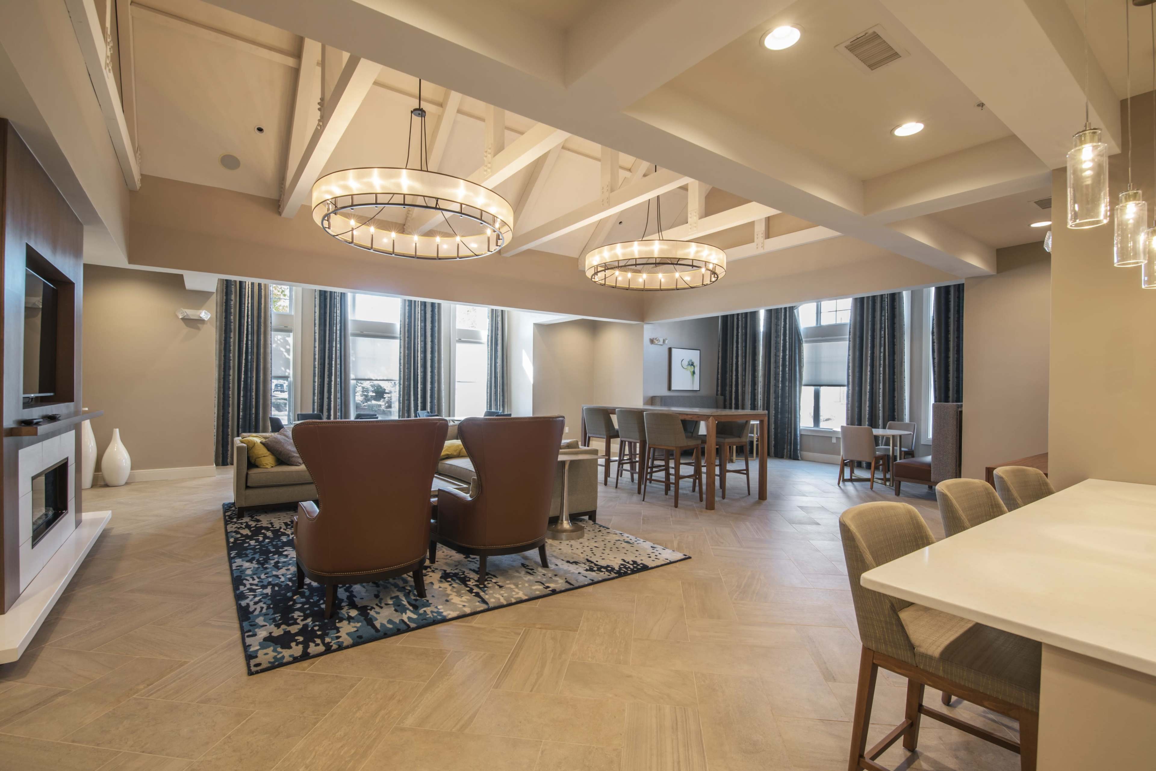 The image shows a modern, open living space with two brown chairs, a large table, and a TV, all under ceiling beams and featuring large windows.
