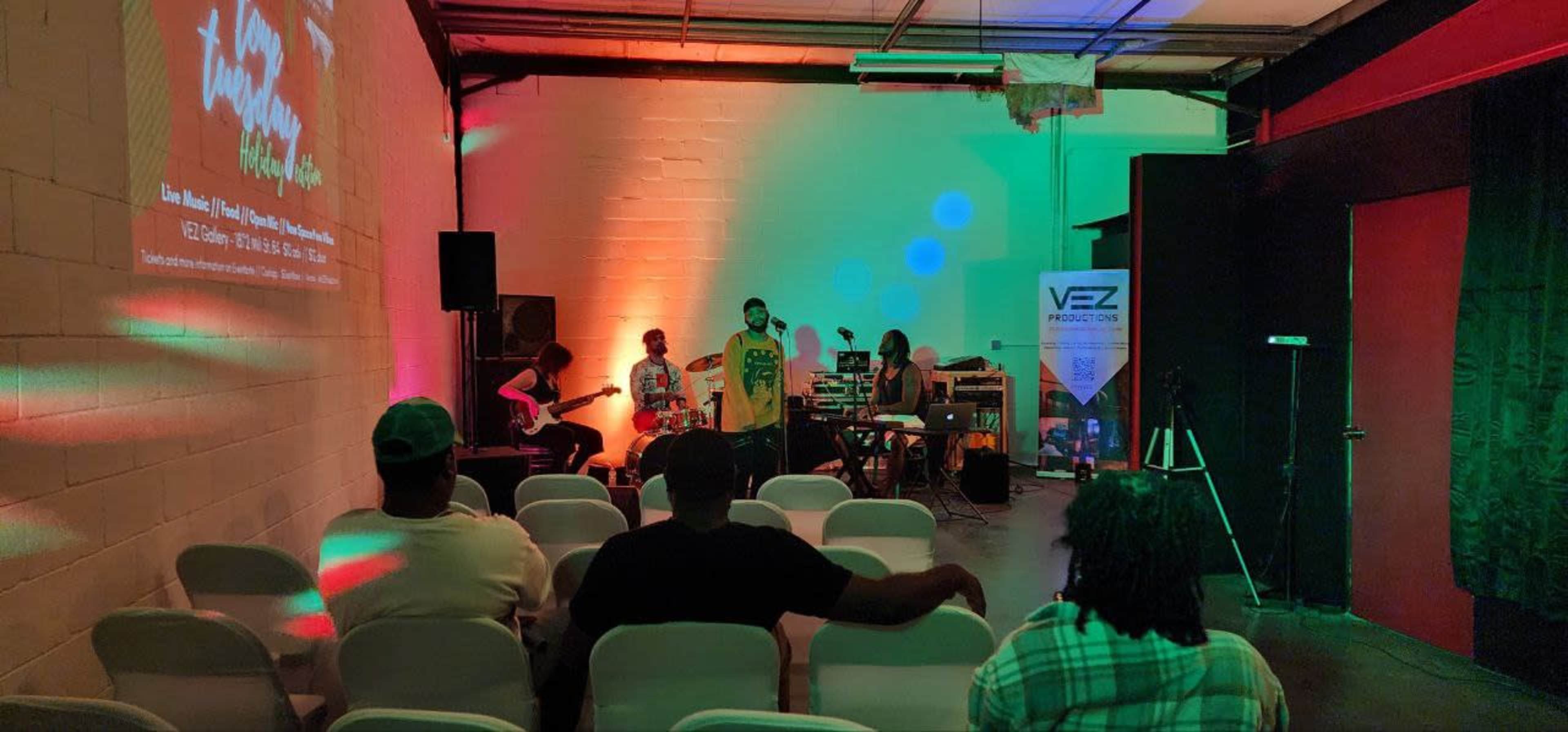 A small audience watches a live music performance in a dimly lit venue with colorful lights and a projector displaying a graphic on the wall.