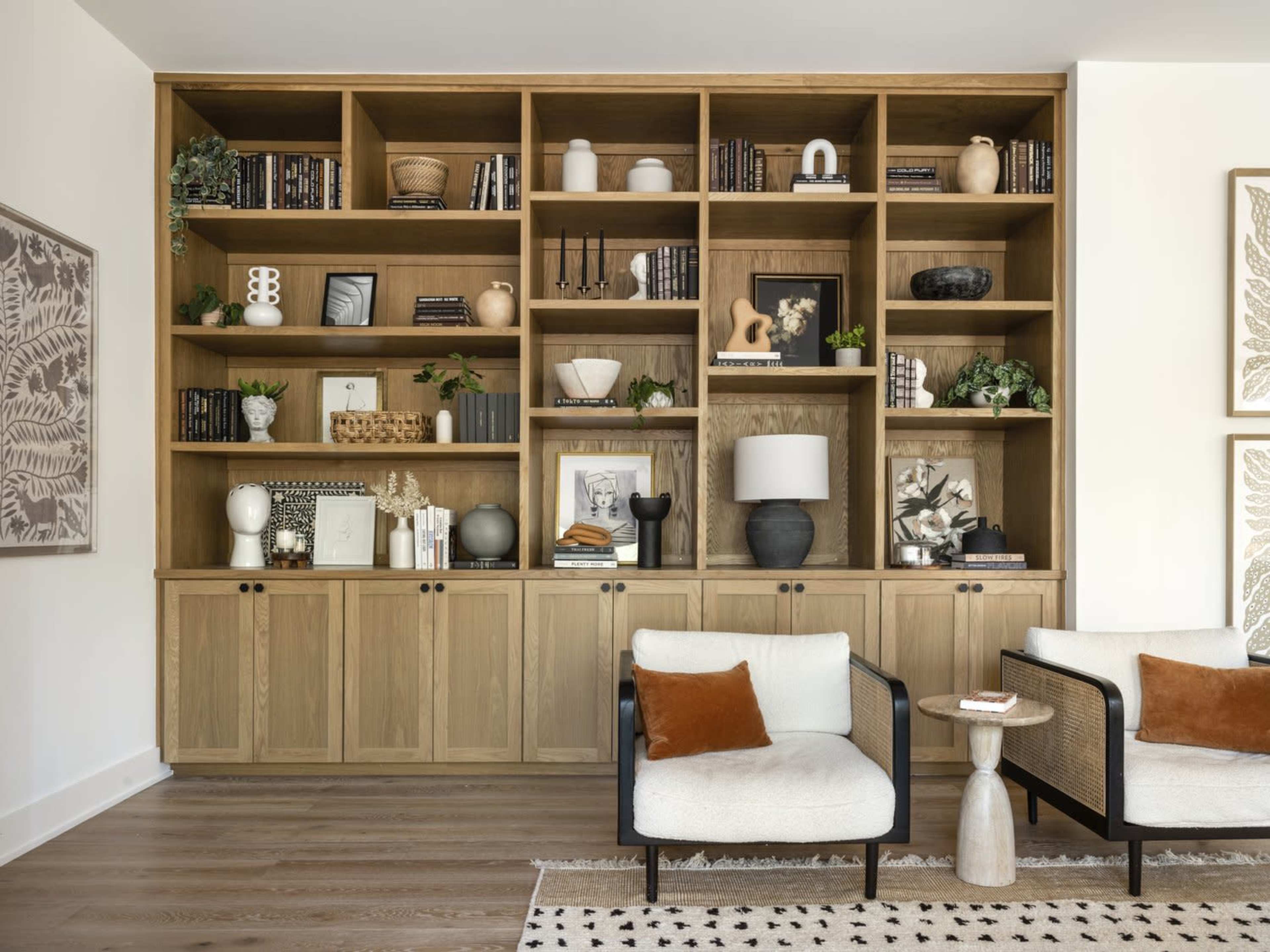 A large wooden bookshelf filled with decorative objects, books, and plants is positioned against a wall, accompanied by two armchairs and a patterned area rug.