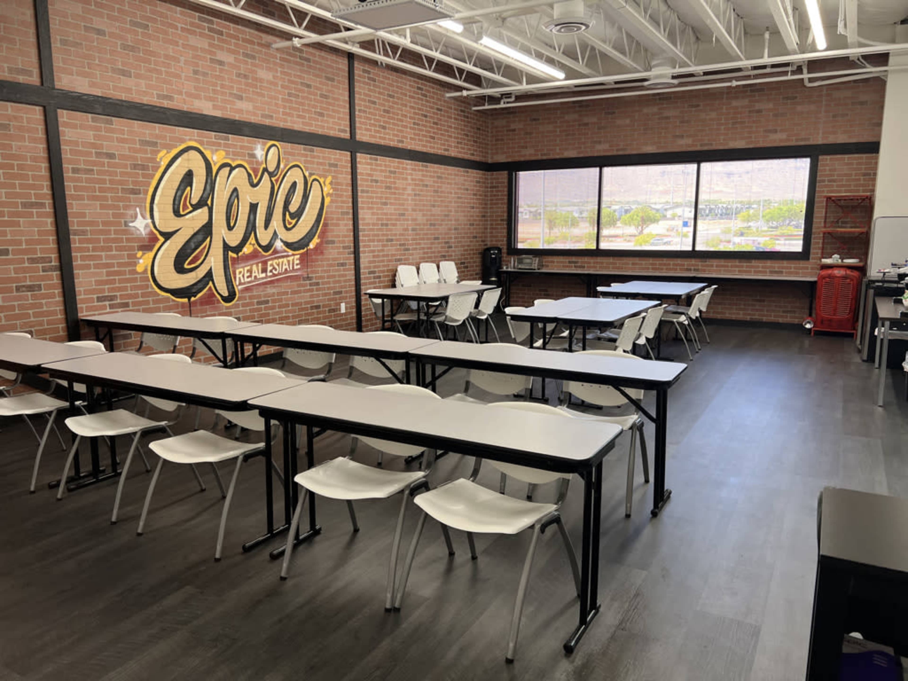 A classroom-style space with white tables and chairs arranged in rows against a brick wall featuring a mural that reads "Epic Real Estate."