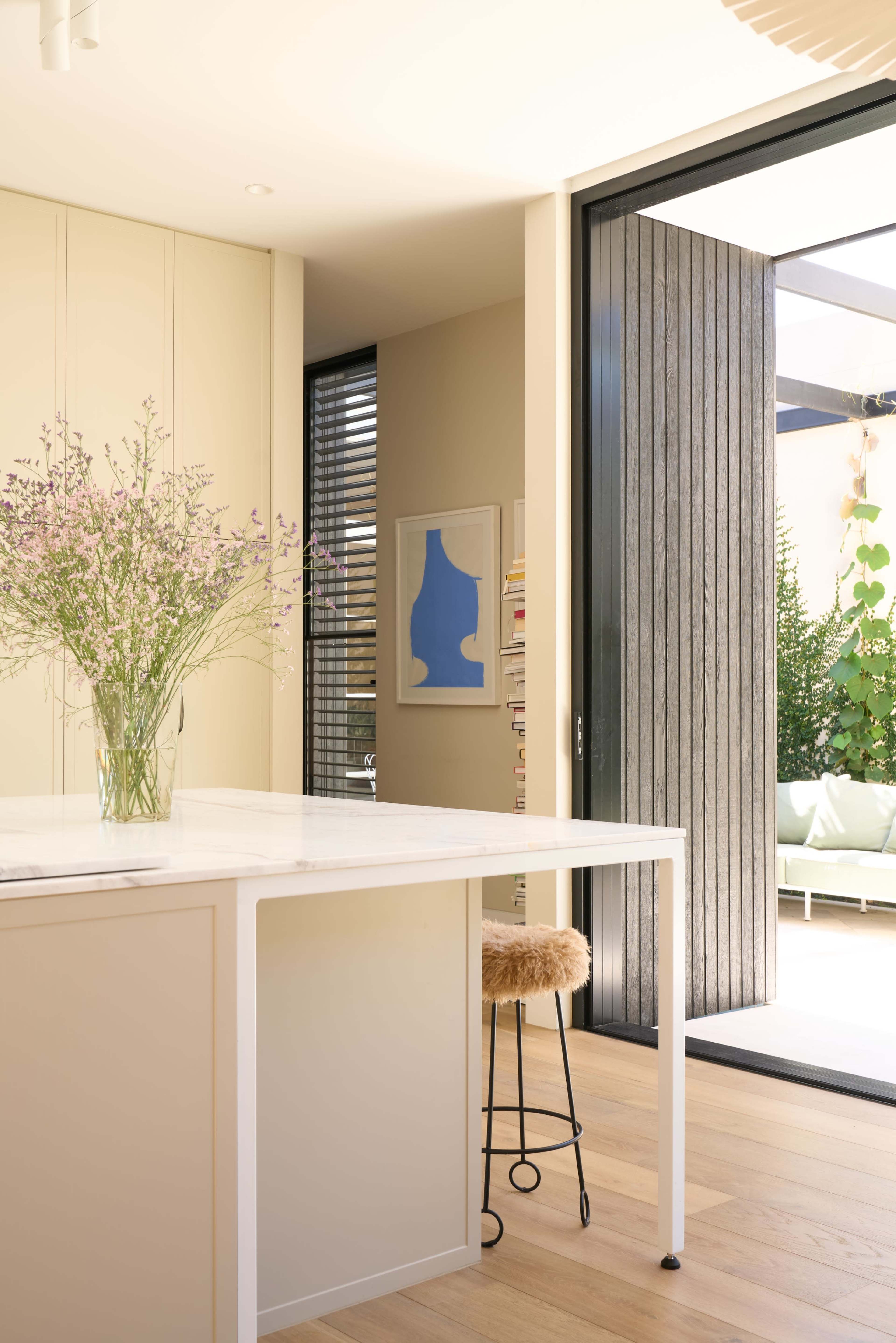 A modern kitchen features a marble island with a vase of flowers and an open door leading to an outdoor space filled with greenery.