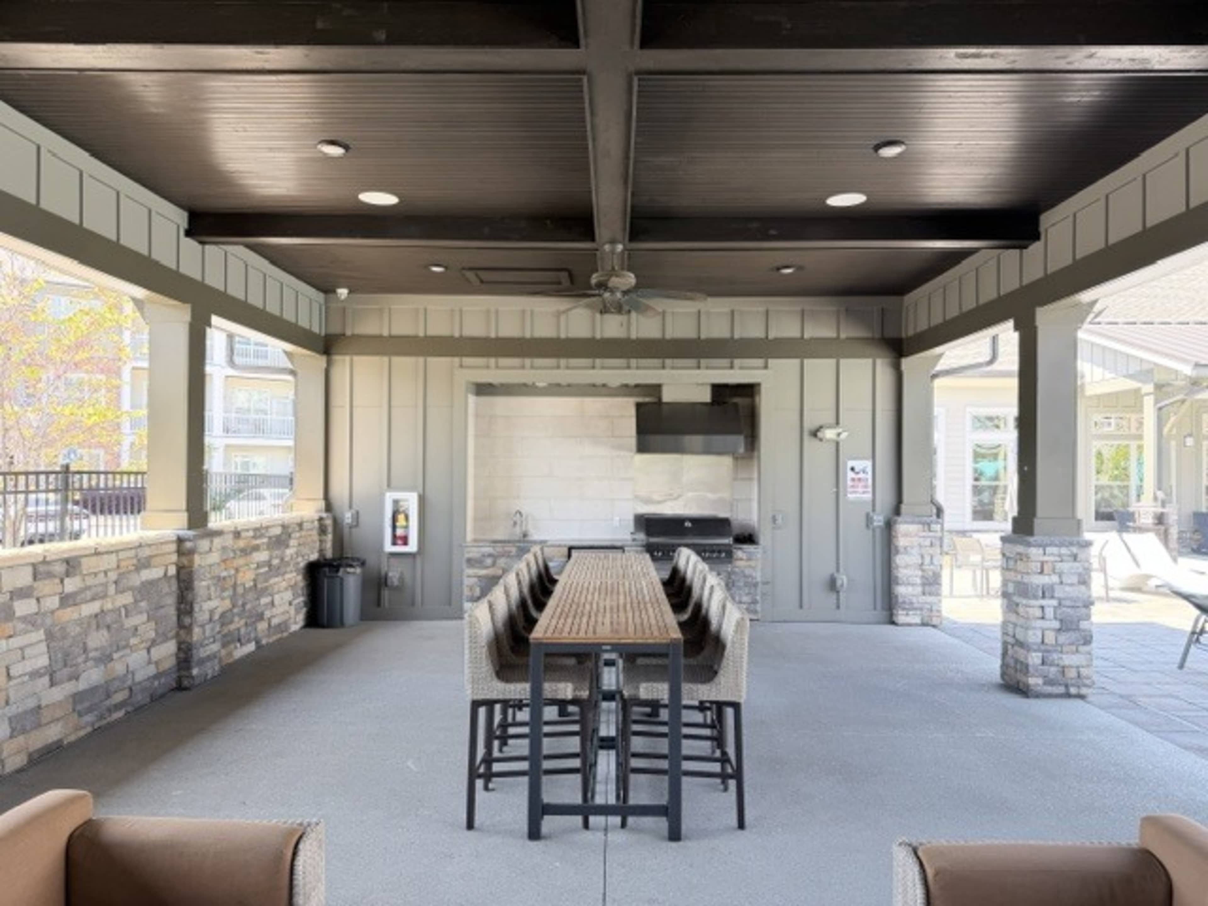 The image shows a covered outdoor seating area with a long wooden table and a grill in the background.