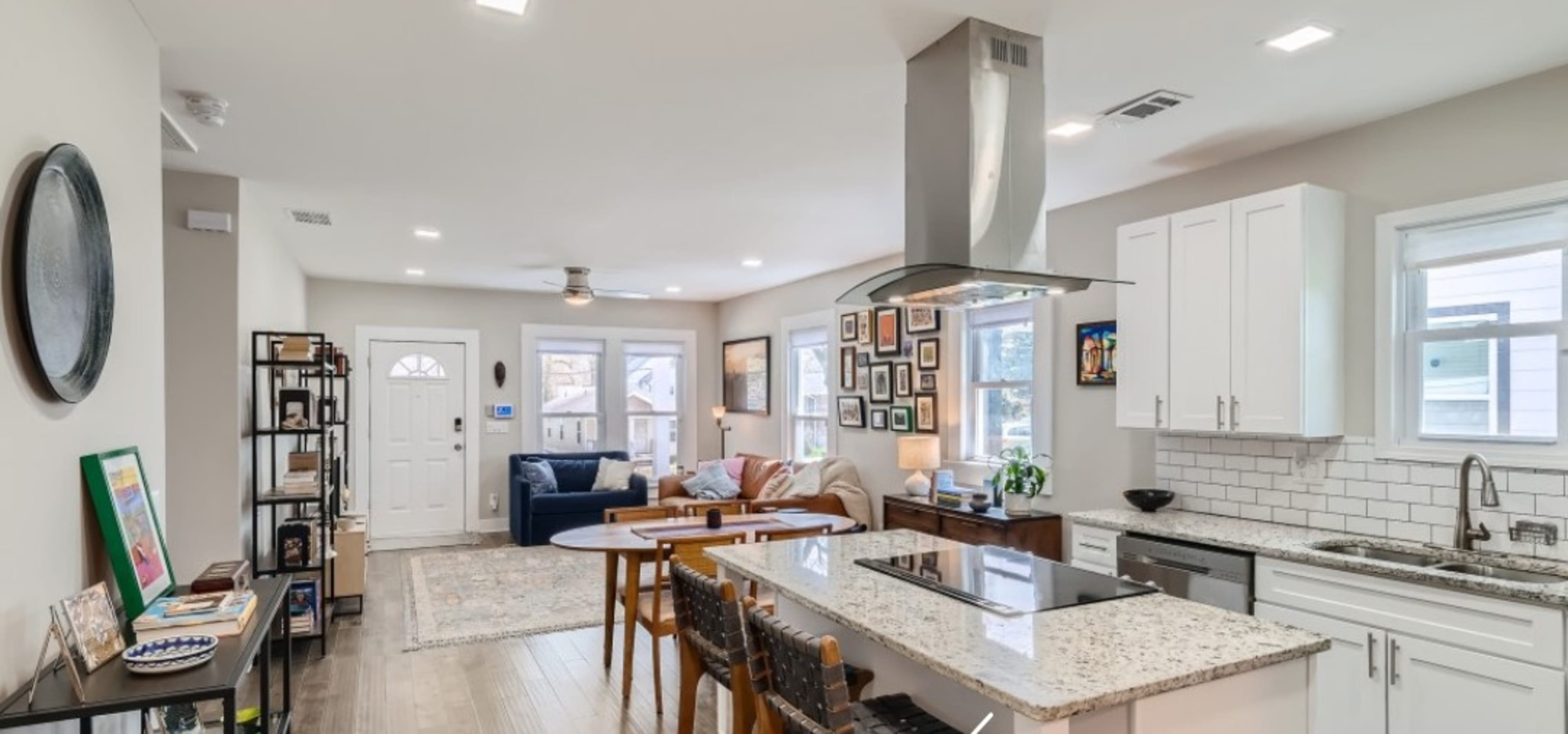 The image shows a modern open-concept living space featuring a kitchen with a breakfast bar, a seating area with a couch, and a front door visible in the background.