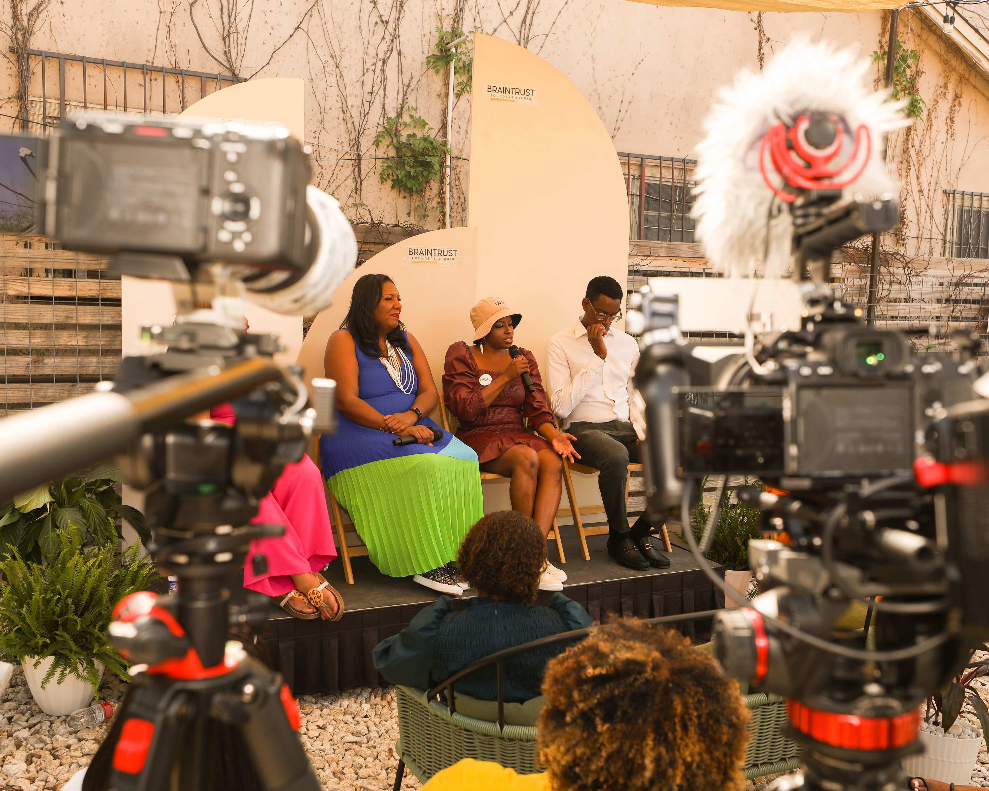 Three speakers sit on a stage during a panel discussion, with cameras focused on them in a garden setting.