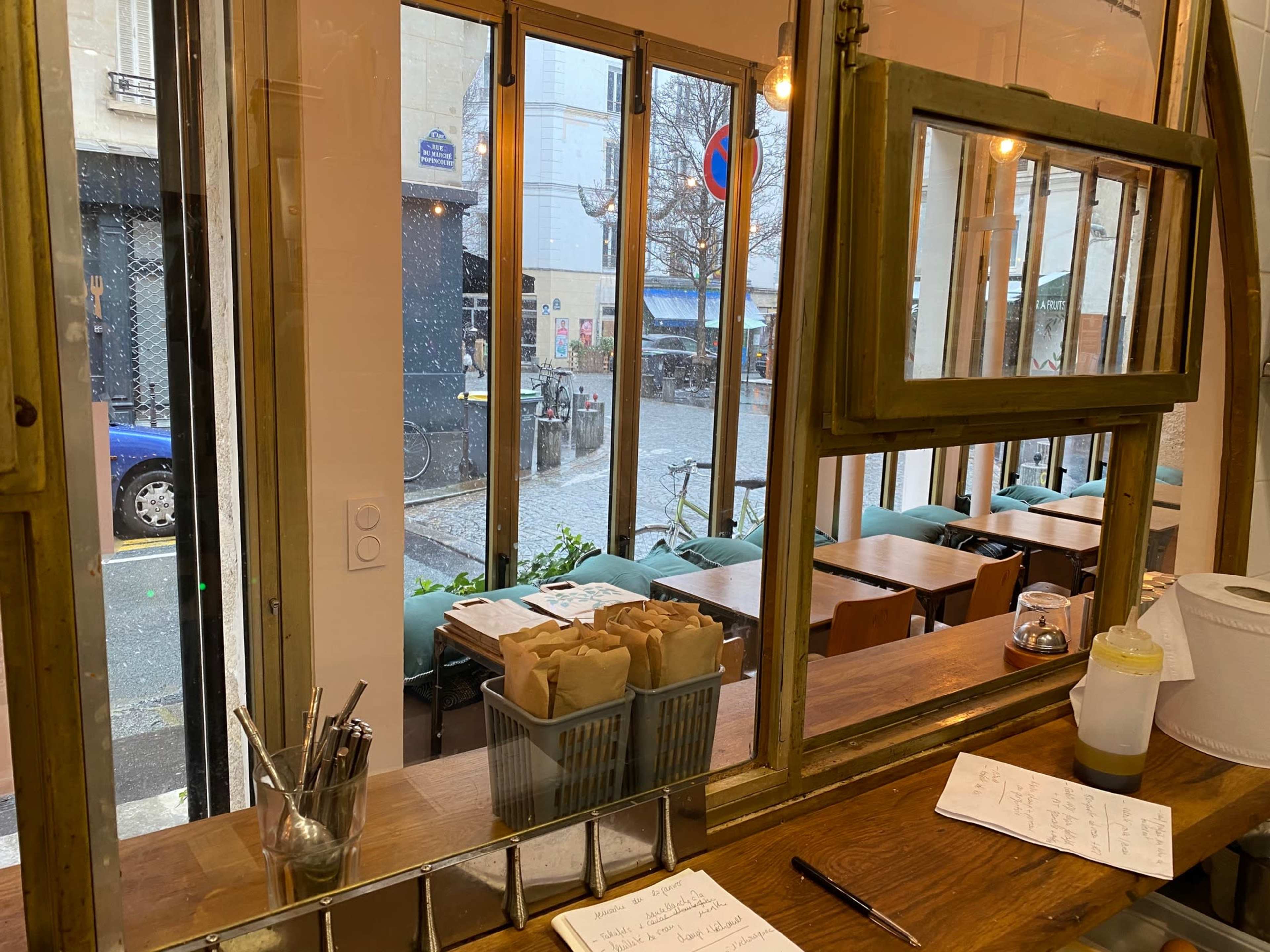 A view of a café interior shows wooden tables and chairs, with large windows displaying a street scene and light rain outside.