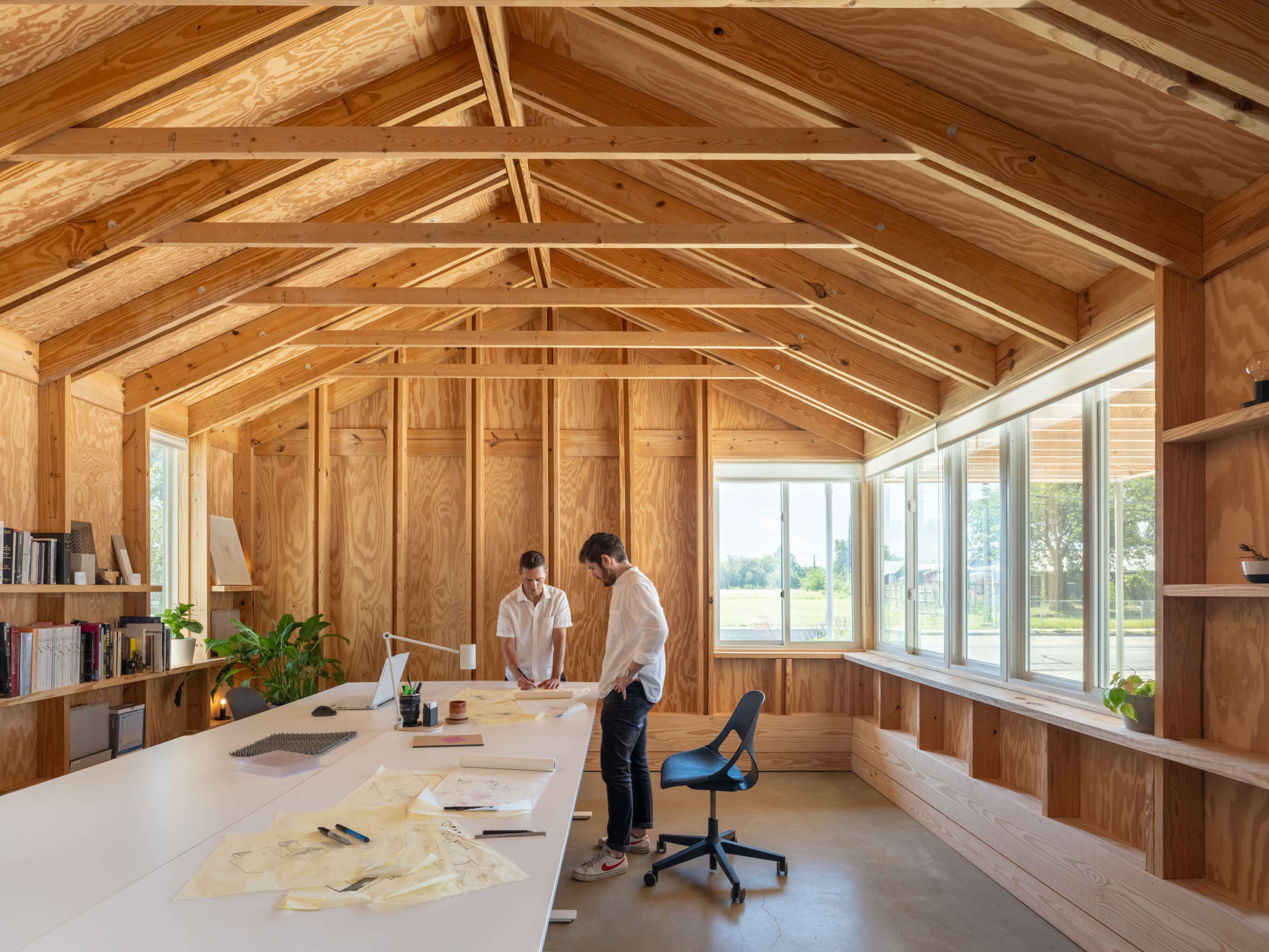 Two individuals stand at a large table in a wooden interior office, examining documents and plans, while natural light streams through large windows.