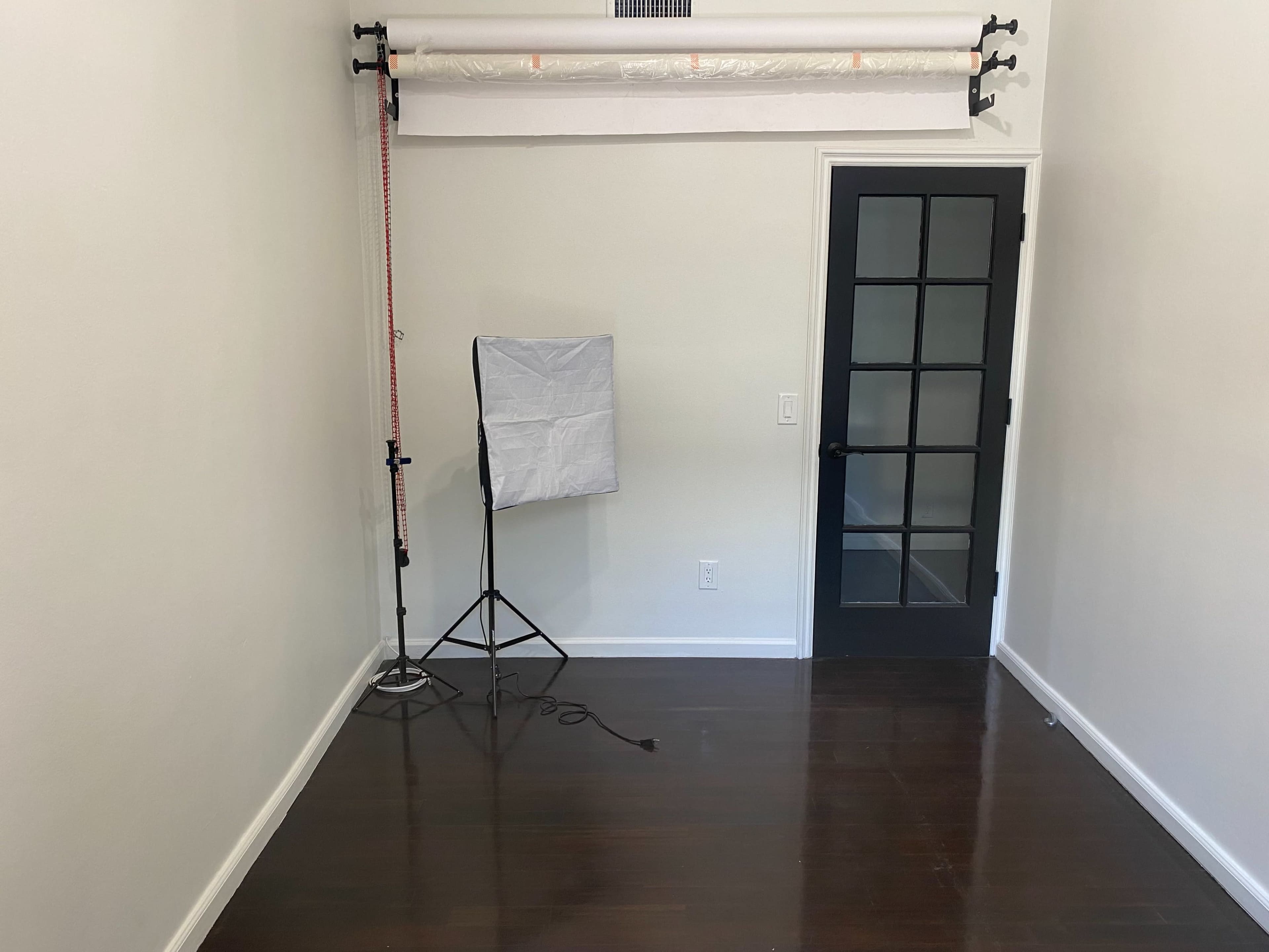 The image depicts a small, sparsely furnished room with a black door, a light stand, and a backdrop set up against one wall.