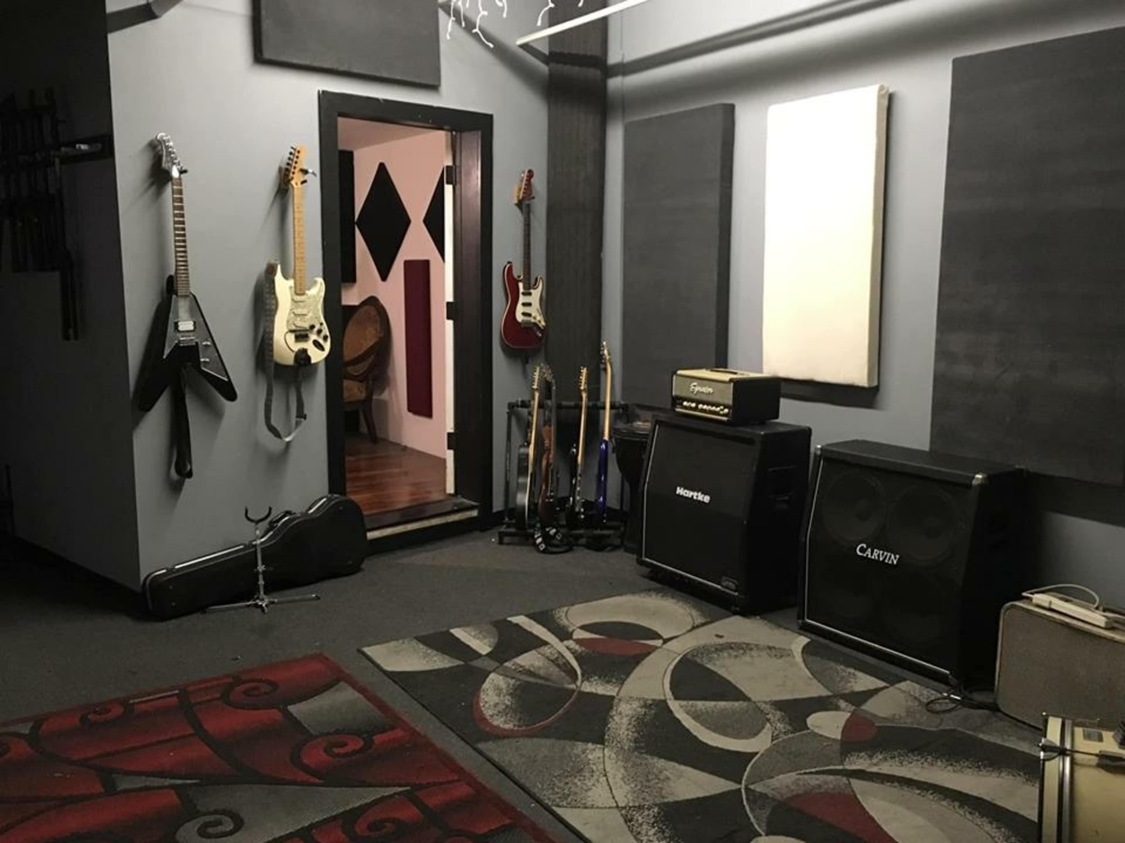 The image shows a music rehearsal room with electric guitars mounted on the wall, amplifiers positioned on the floor, and a patterned area rug.