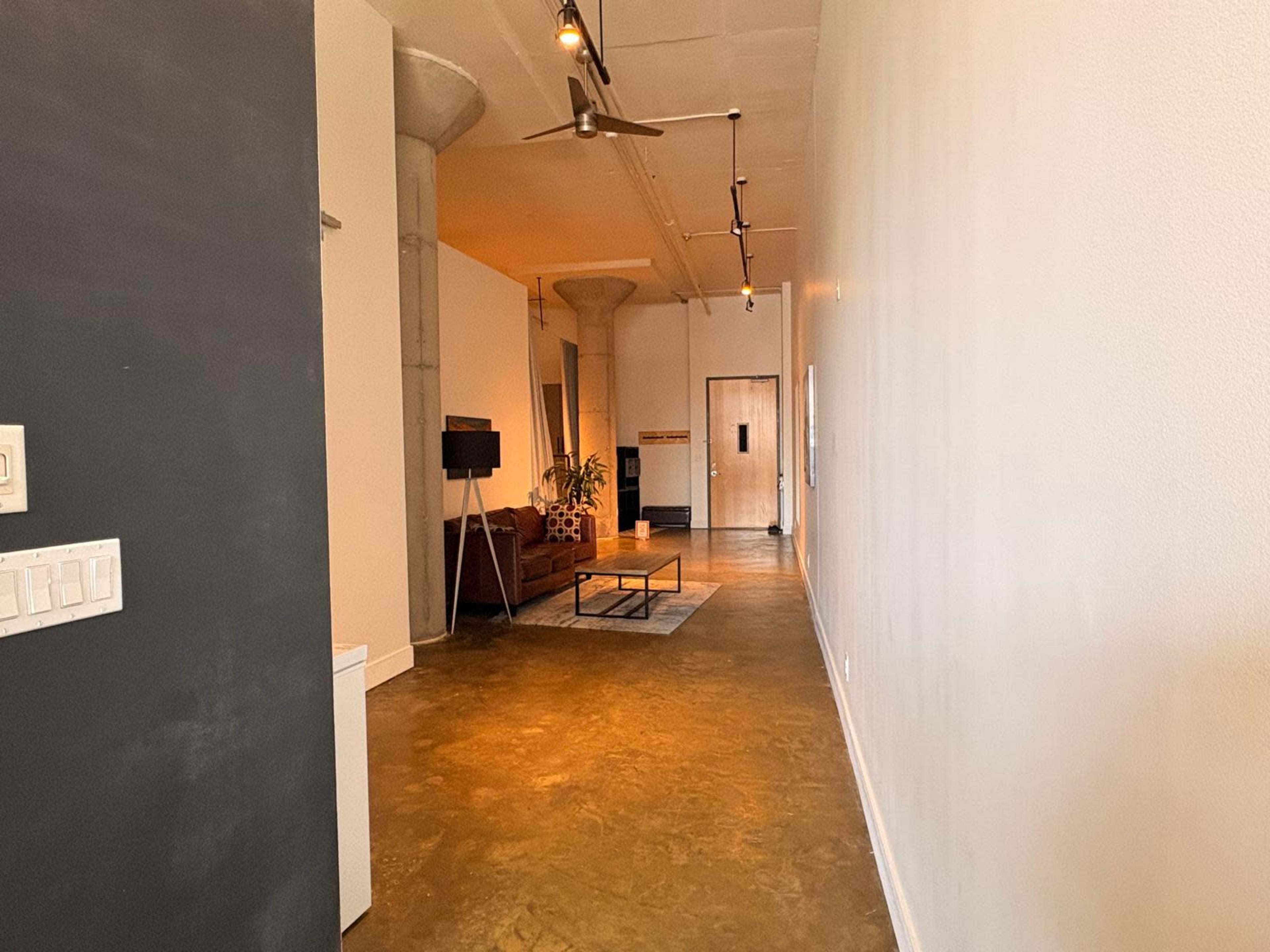 The image depicts a long, narrow hallway with a polished concrete floor, featuring a couch, a coffee table, and a plant at the far end.
