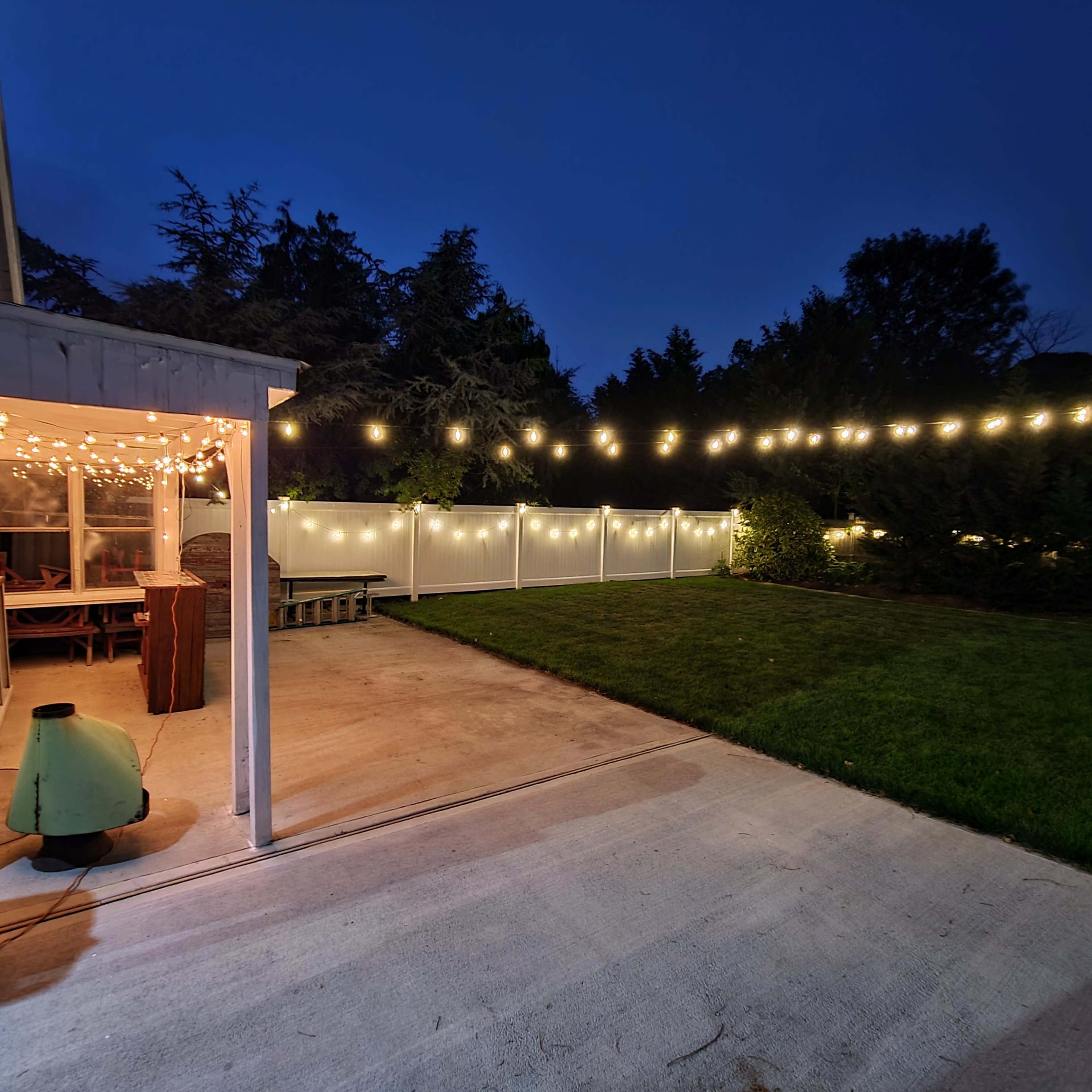 The image shows a backyard with string lights illuminating a grassy area alongside a concrete patio and a white fence.