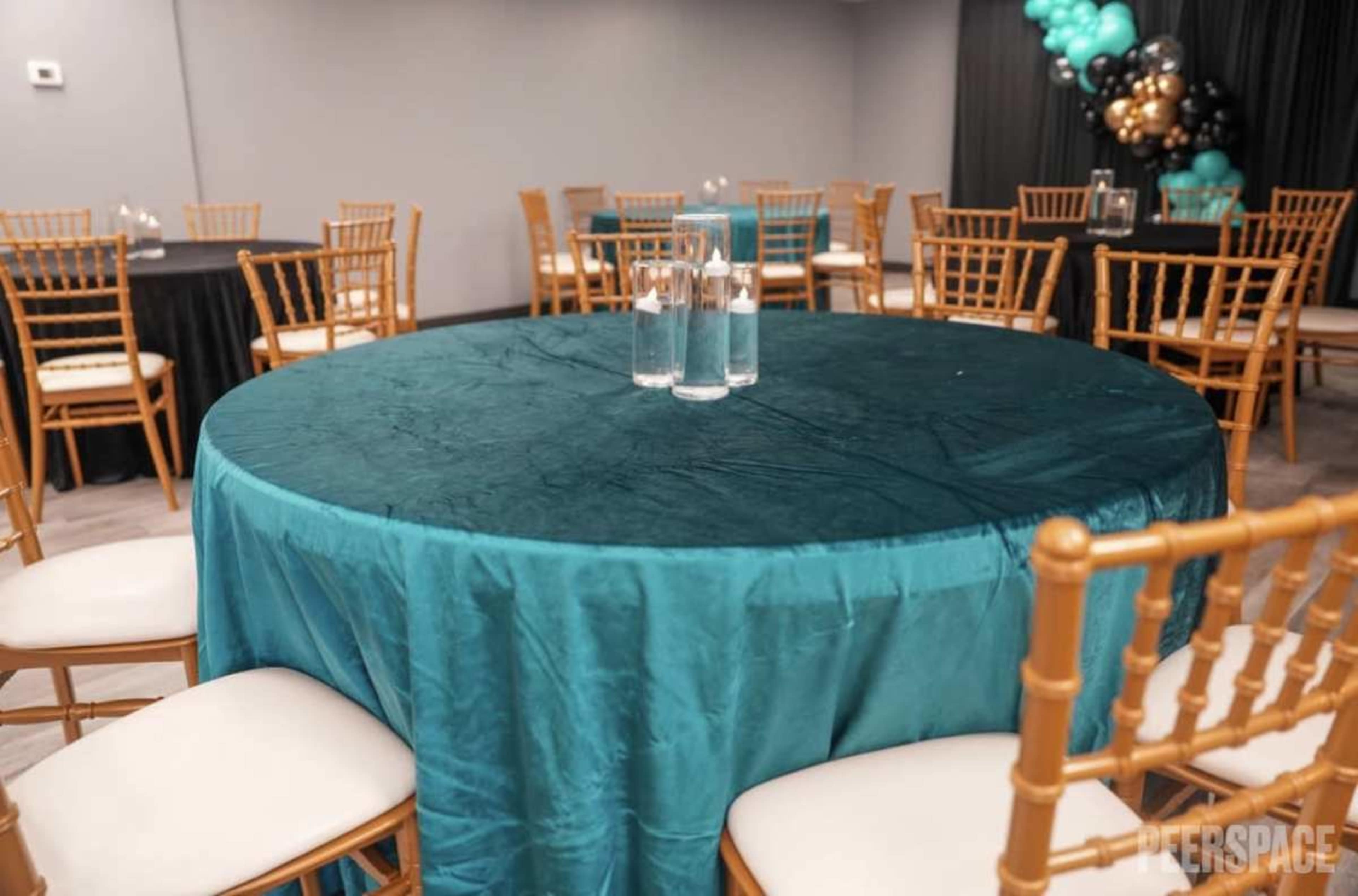 A round table covered with a teal tablecloth features three glasses of water in the center, surrounded by gold-framed chairs in a banquet setting.
