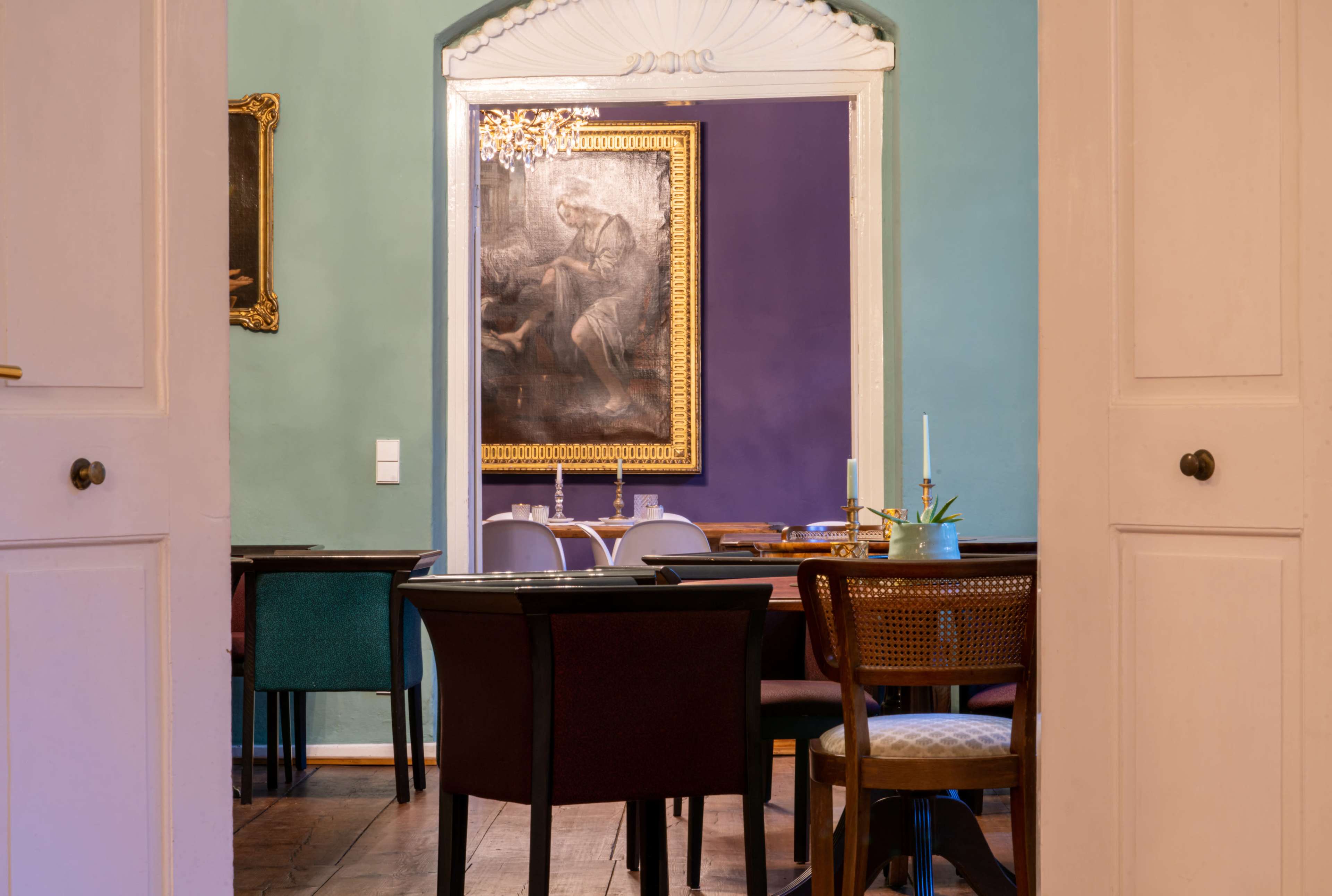 A dining area with various chairs around tables, framed artwork on the wall, and a doorway leading to another room.