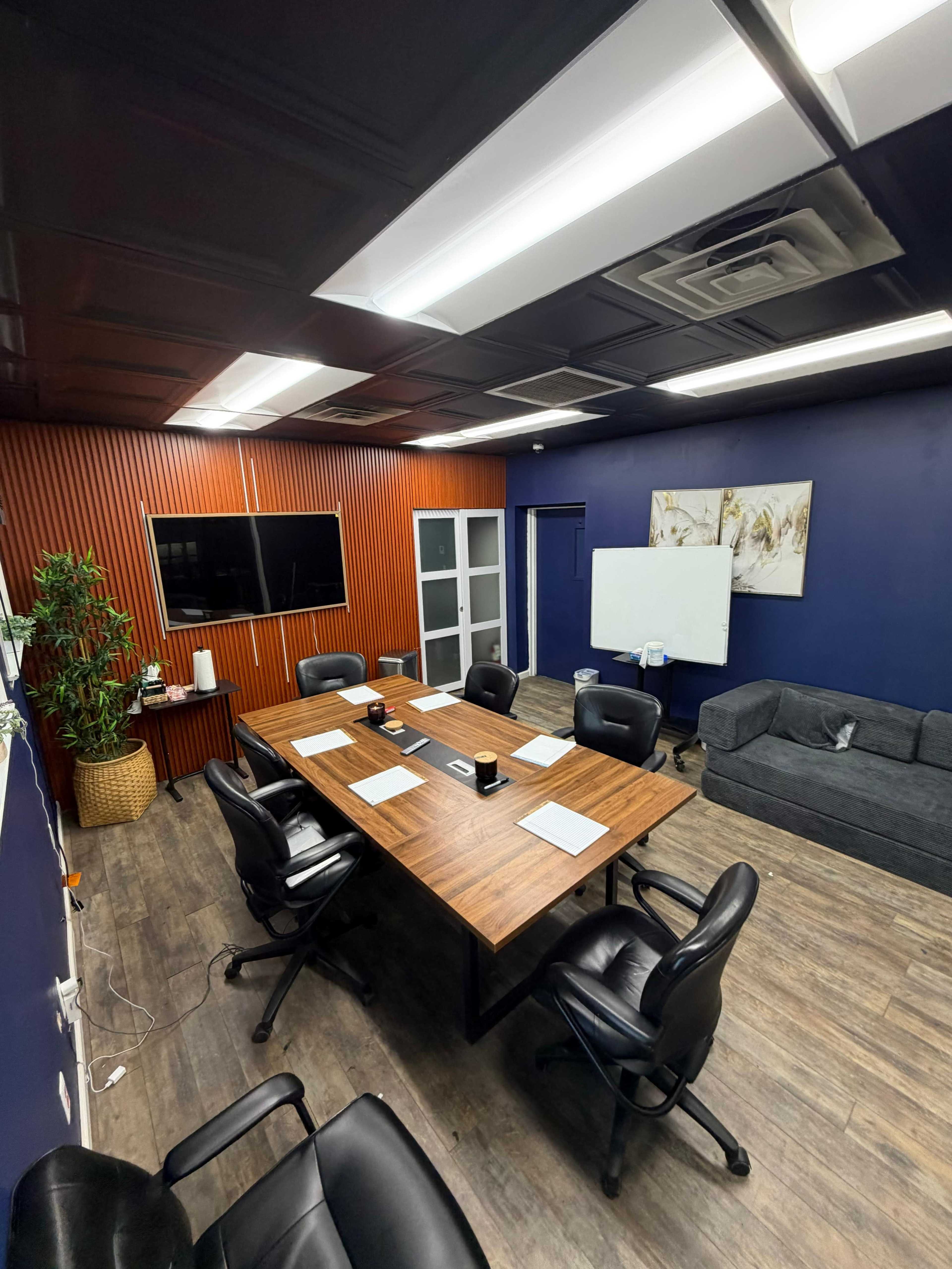 A modern conference room features a large wooden table surrounded by black chairs, a couch, a whiteboard, and a wall-mounted screen.