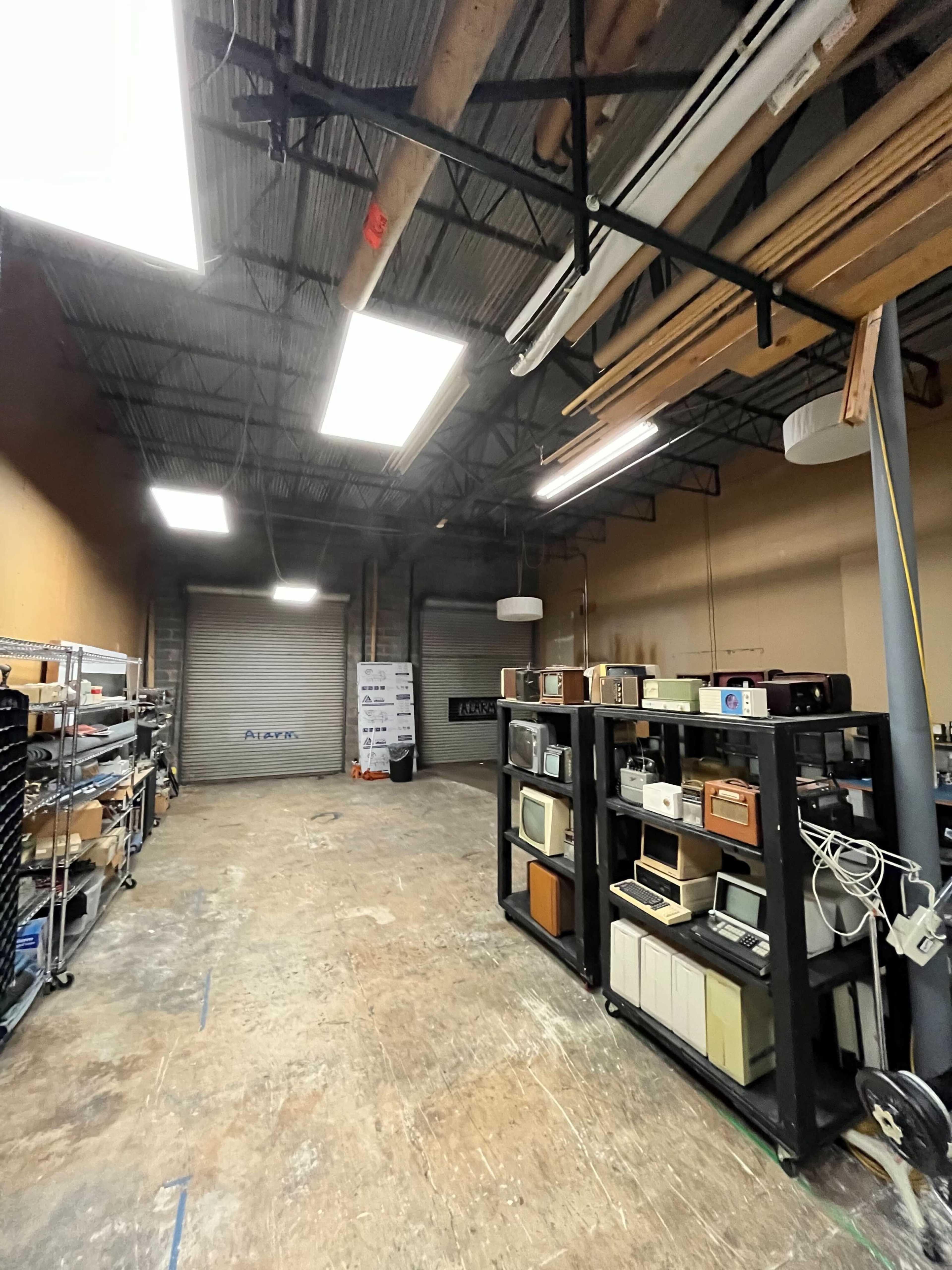 The image shows a spacious industrial room with shelves containing various electronic devices and items, and two roll-up doors at the far end.