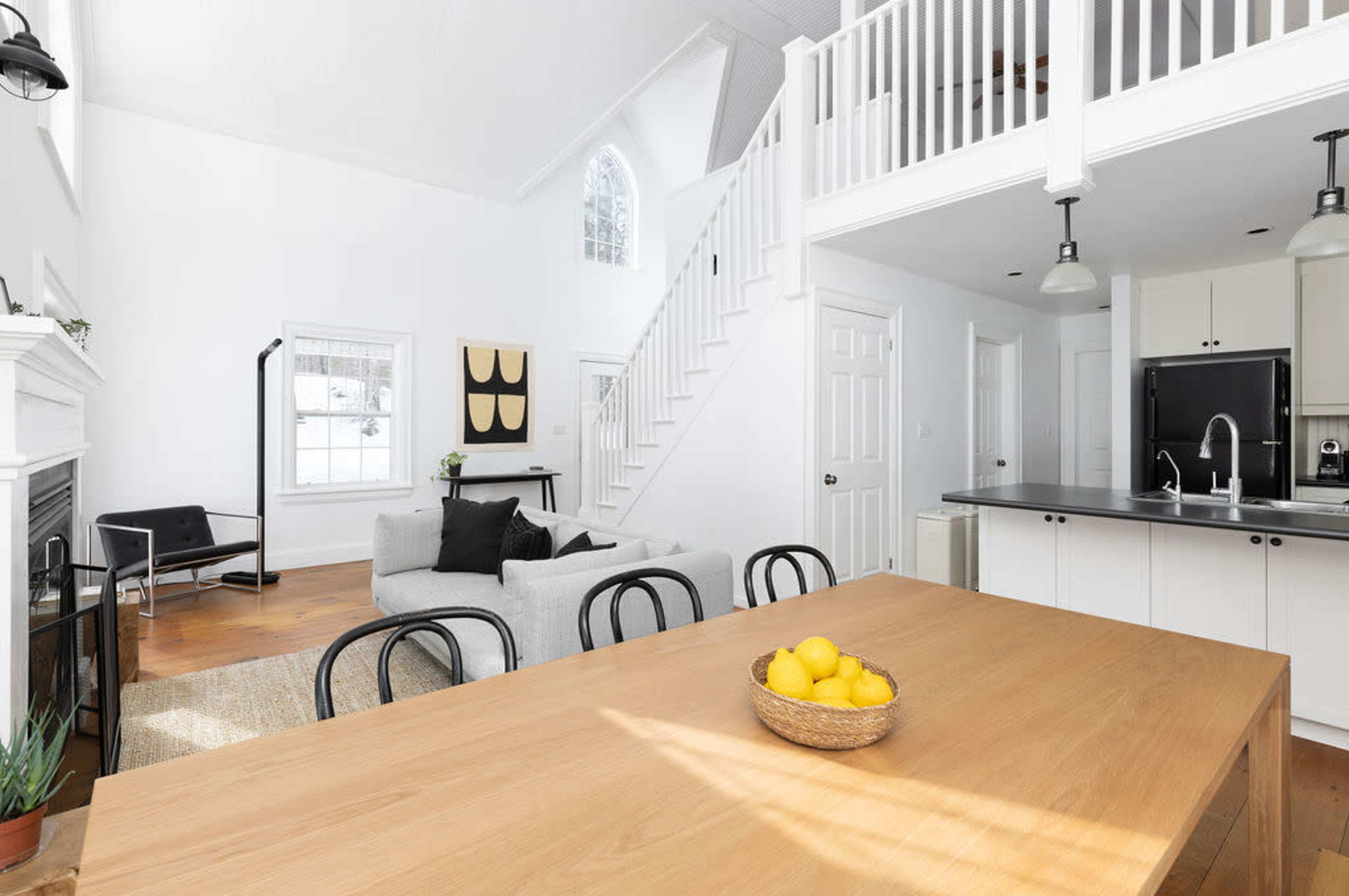 The image shows a bright, modern living space with a high ceiling, featuring a kitchen area, dining table with a bowl of lemons, a gray sofa, and a staircase leading to a loft.