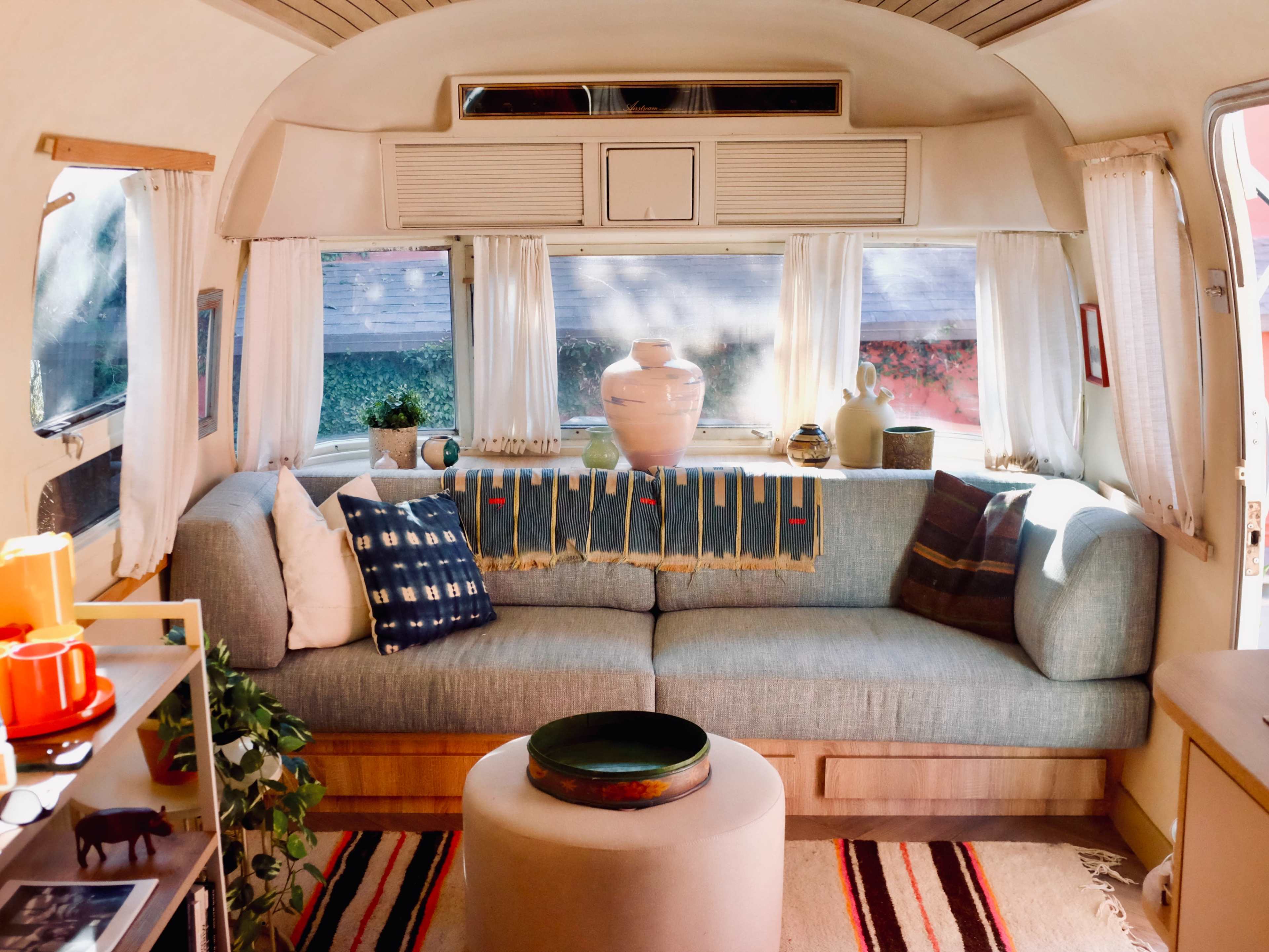 The image shows a cozy interior of an Airstream trailer with a sofa, decorative pillows, a small table, and large windows allowing natural light to enter.